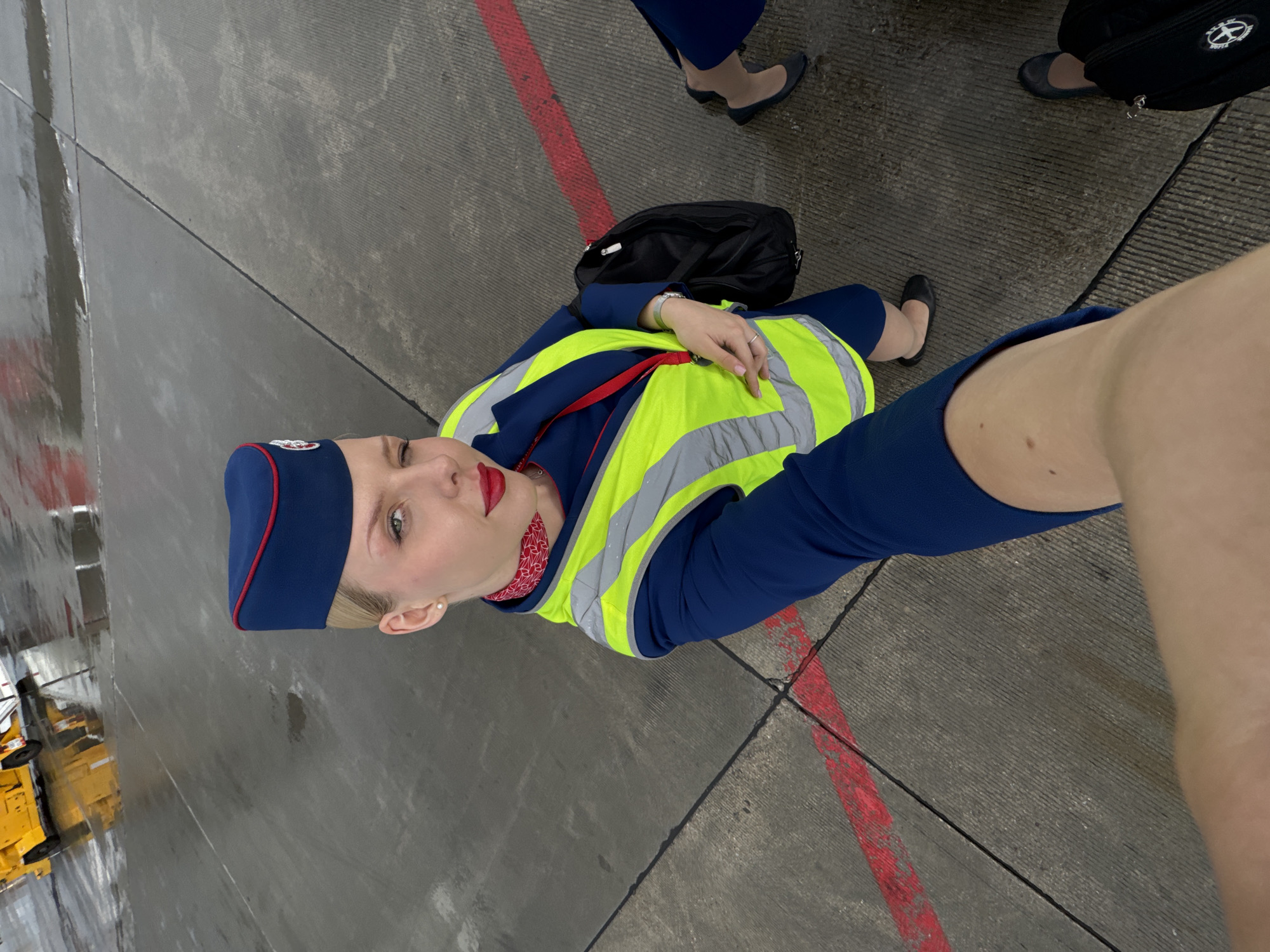 A female flight attendant stands on an airport tarmac. She wears a navy blue uniform with a matching hat, a red and white patterned scarf, and a bright yellow safety vest with reflective stripes. Her makeup is immaculate, with bold red lipstick. She has a confident and friendly expression. A black duffel bag is visible behind her. In the background, another person in a similar uniform can be seen, along with part of a yellow vehicle, possibly airport ground equipment. The tarmac is wet, suggesting recent rain or mist, and the lighting indicates it is daytime. The overall scene depicts a professional at work in an airport environment.