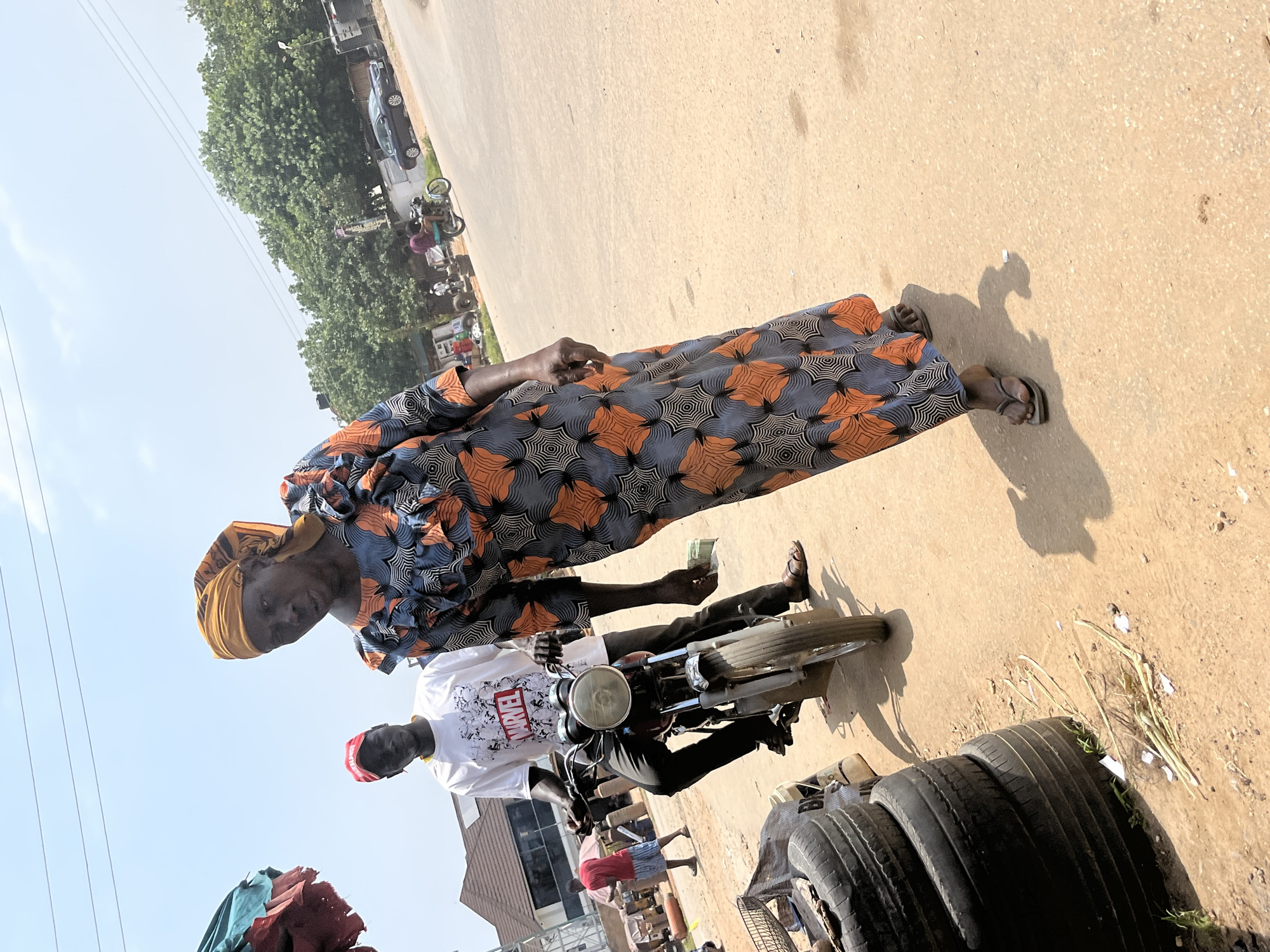 In an outdoor scene in Abuja, Nigeria, a woman in a colorful, long, patterned dress and a yellow headscarf walks towards the camera on a dusty, sunlit street. To her left, a young man wearing a white Marvel t-shirt and a red cap rides a motorcycle. Behind them, other people and vehicles are visible, including a motorcycle with two riders in the distance, and parked cars. The environment includes buildings, trees, and a stack of old tires. The sky is bright with some clouds.