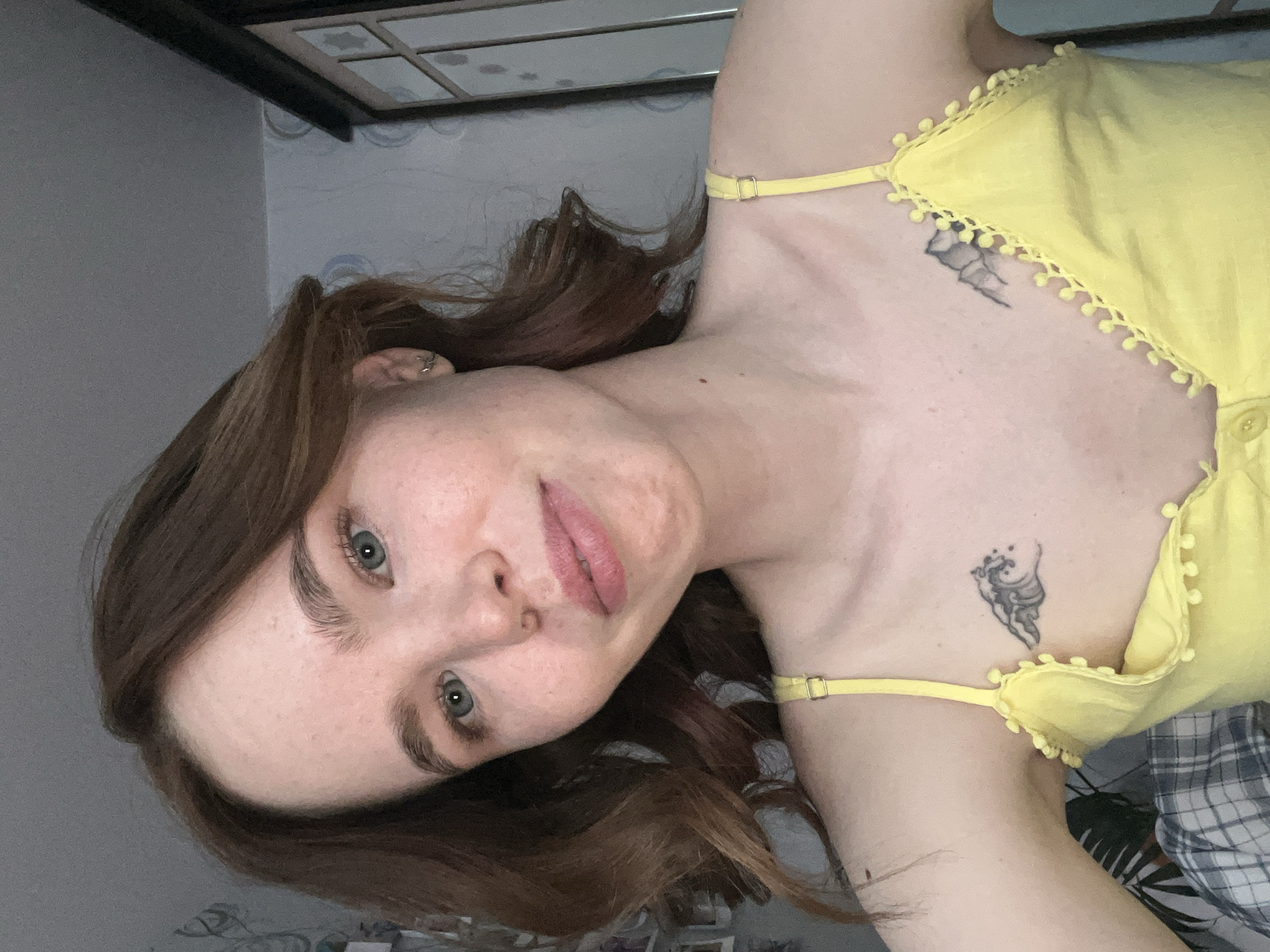 This image depicts a close-up, head-and-torso view of a young adult female positioned indoors in the city of Chaikovsky, Russia. The subject has light skin with visible freckles, reddish-brown wavy hair, and light-colored eyes. She is looking directly at the camera with a faint smile. She wears a yellow spaghetti-strap top adorned with small spherical embellishments along the neckline. A small silver hoop earring is visible in her left ear. Two black and grey tattoos are present on her upper chest: a stylized wave on her anatomical left side and a mountain range on her anatomical right side. The background reveals patterned wallpaper with light blue circular designs, partially obscured framed items on the wall to the left, and a dark multi-paneled cabinet or wardrobe to the right. A plaid fabric is visible at the bottom left of the frame. The ceiling above is plain grey.