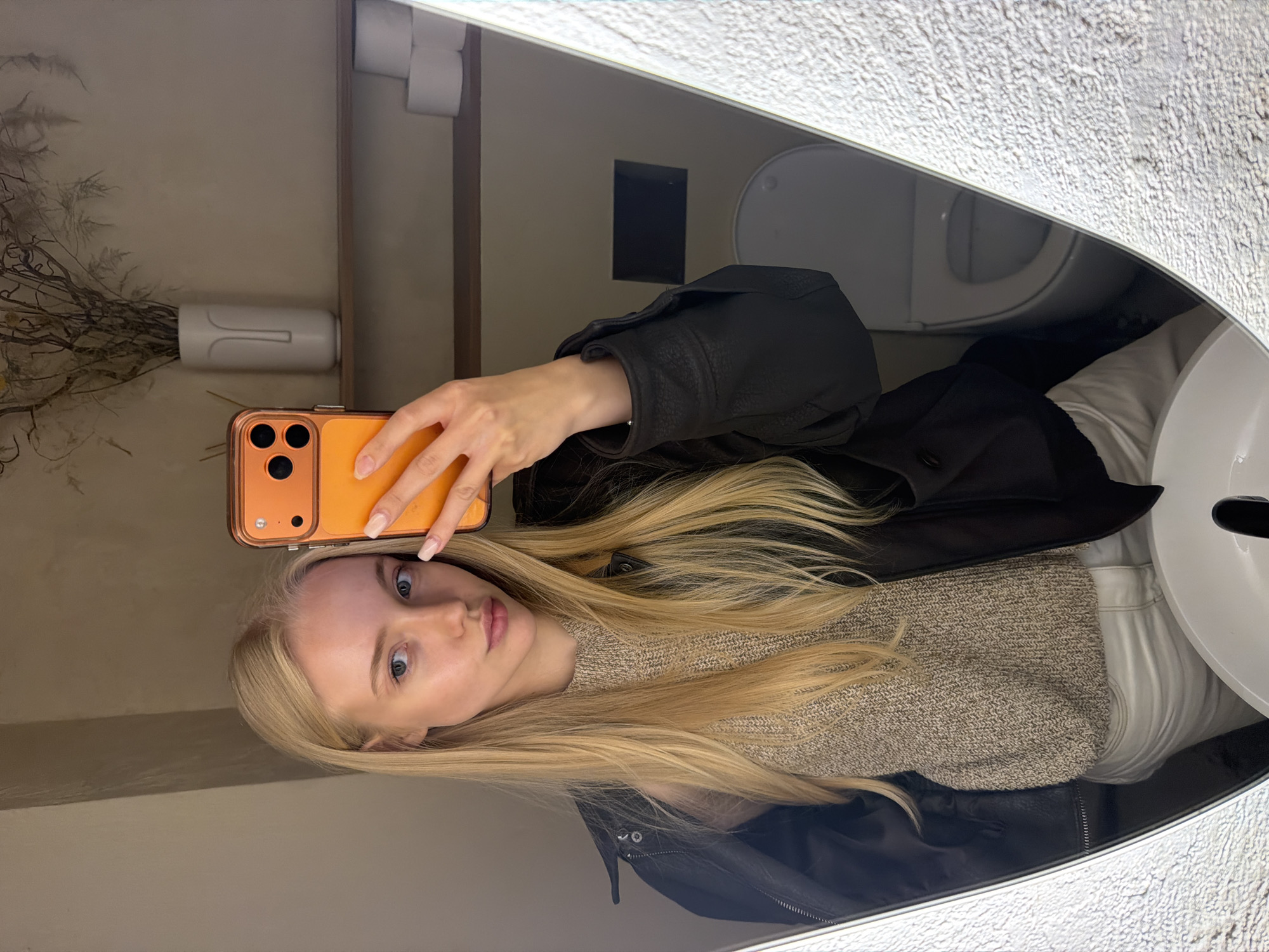 A young blonde female is captured in a mirror selfie within a bathroom setting, located in Unknown, Unknown. She has long, straight, blonde hair and is wearing a light beige knit sweater under a dark (likely black) jacket draped over her shoulders. Light-colored pants are partially visible below the sweater. Her right hand holds an orange-cased smartphone, with its back facing the viewer, positioned to take the photograph in the mirror. Her fingernails have a light, neutral polish. The mirror itself has an irregular, rounded shape and appears to have a textured or illuminated frame on its vertical edges. Reflected in the mirror, the background reveals a light beige wall. A wooden shelf is mounted on the wall, displaying a white, minimalist vase holding dried botanical elements, including twigs and grasses. To the right of the vase, three rolls of white toilet paper are stacked on the shelf. The rim of a white sink basin is visible in the lower center foreground of the reflection, while a white toilet, with its lid open, is partially seen in the lower right background.
