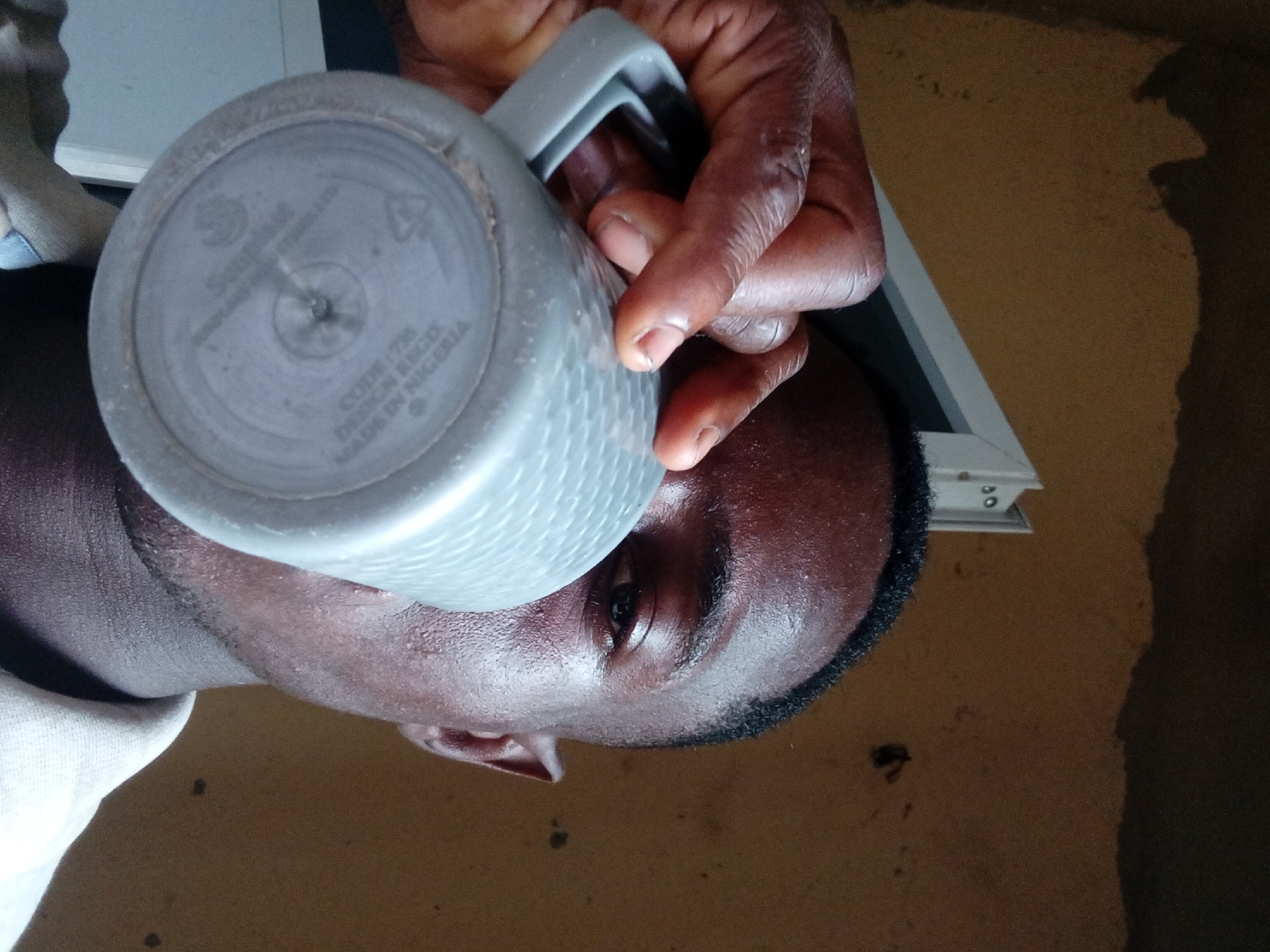 A person with dark skin is holding a gray mug to their face. The mug has a circular bottom and handle. The text "MADE IN NIGERIA" is visible on the bottom of the mug. The person is indoors, and a wall or surface is visible in the background. The location is Akwete, Nigeria.