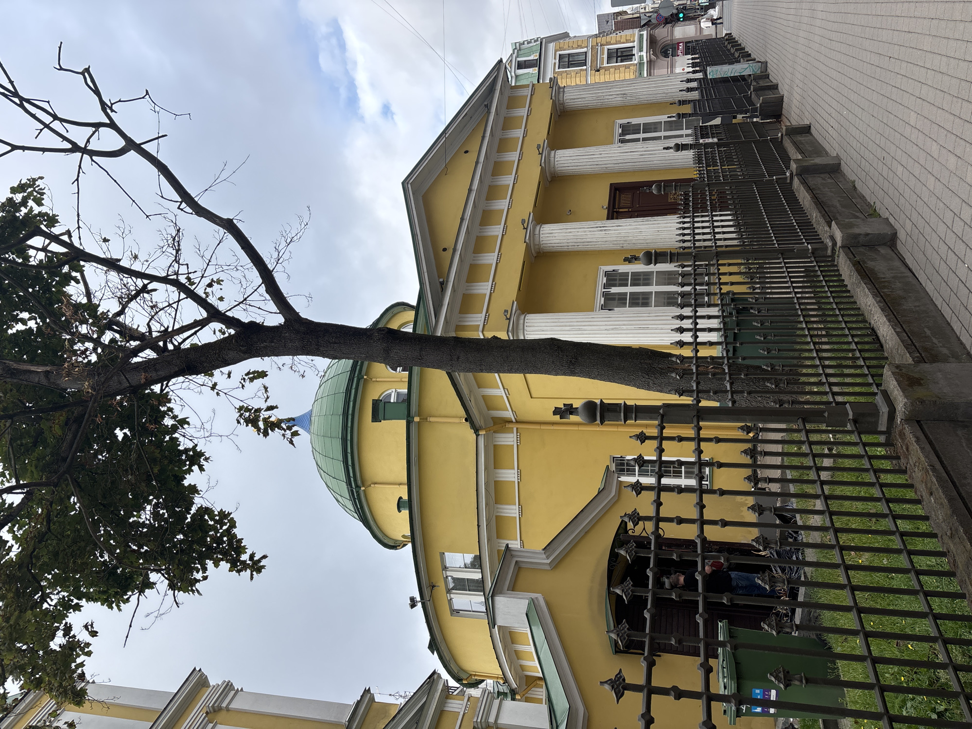A yellow building with a green domed roof and white columns is shown in Riga, Latvia. A tree with sparse foliage stands in front of the building. A metal fence separates the building from a paved sidewalk. A green trash can is visible next to the entrance of the building. A person wearing a black shirt and blue jeans is standing in the doorway. A few black trash bags are placed near the entrance. The sky is overcast with some clouds. The building appears to be a church or a public institution.