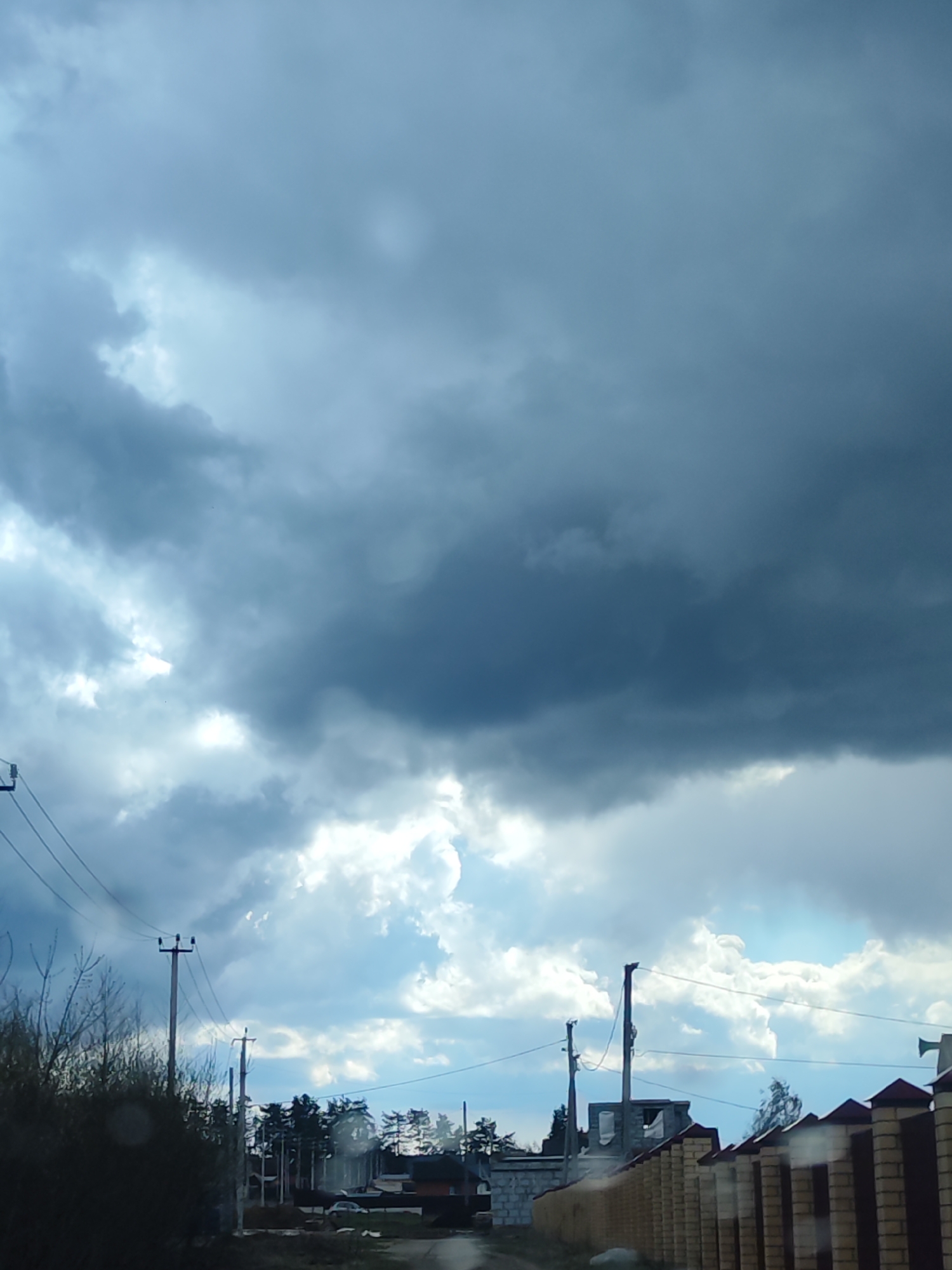 The image captures a rural or semi-urban landscape under a dramatic, overcast sky. The majority of the frame is dominated by heavy, dark grey storm clouds, interspersed with brighter patches of white and light grey clouds where sunlight appears to be breaking through. This suggests unsettled weather, possibly before or after rain, during daytime.
In the foreground, on the left, there are sparse, bare tree branches or bushes, indicating a colder season. Several wooden utility poles with numerous power or communication lines extend horizontally across the lower part of the image. On the right, a robust fence with brick pillars and dark panels is prominently featured.
The middle ground reveals more signs of human settlement. Clusters of trees, some appearing coniferous, dot the landscape. Residential structures are visible, including one on the left that appears to be under construction with grey cinder block walls. An unpaved road or path is faintly visible in the bottom center.
No people or animals are present in the scene. There is no discernible text. The overall atmosphere is somewhat moody due to the heavy cloud cover, contrasted by the glimpses of brighter sky. The image portrays a quiet, ordinary day in a residential area of Chaikovsky, Russia, under an impressive sky.