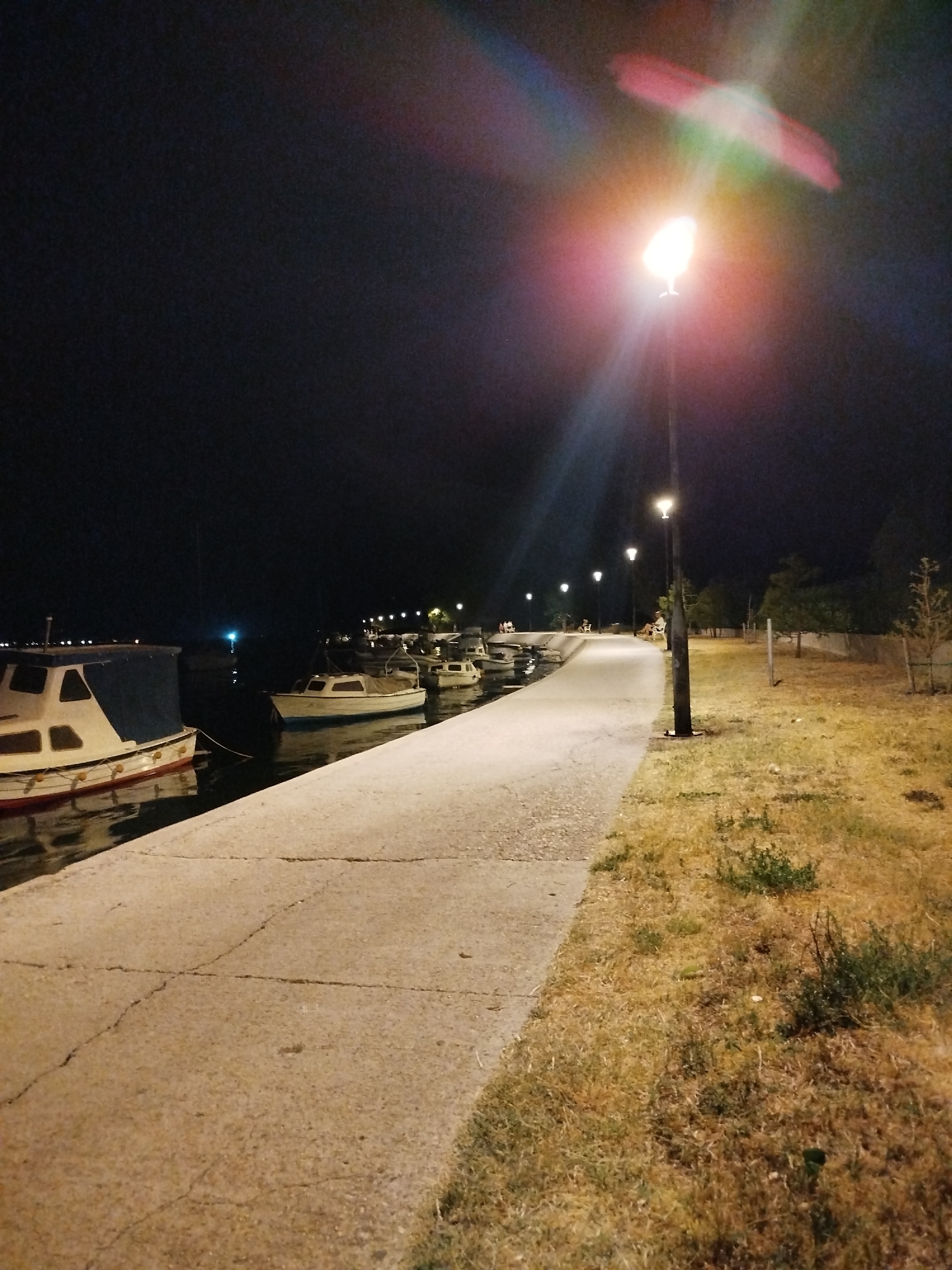 The image depicts a nighttime scene along a waterfront promenade in Omiš, Croatia. A paved walkway curves along the edge of a body of water, where several small boats are moored. The water reflects the light from a street lamp overhead, which casts a bright glow and creates lens flare effects, including streaks of red and green light. Dry, yellowish grass borders the walkway on the right side, with sparse trees and vegetation further in the background. A few indistinct figures can be seen sitting on benches further down the promenade. The overall atmosphere is calm and quiet.