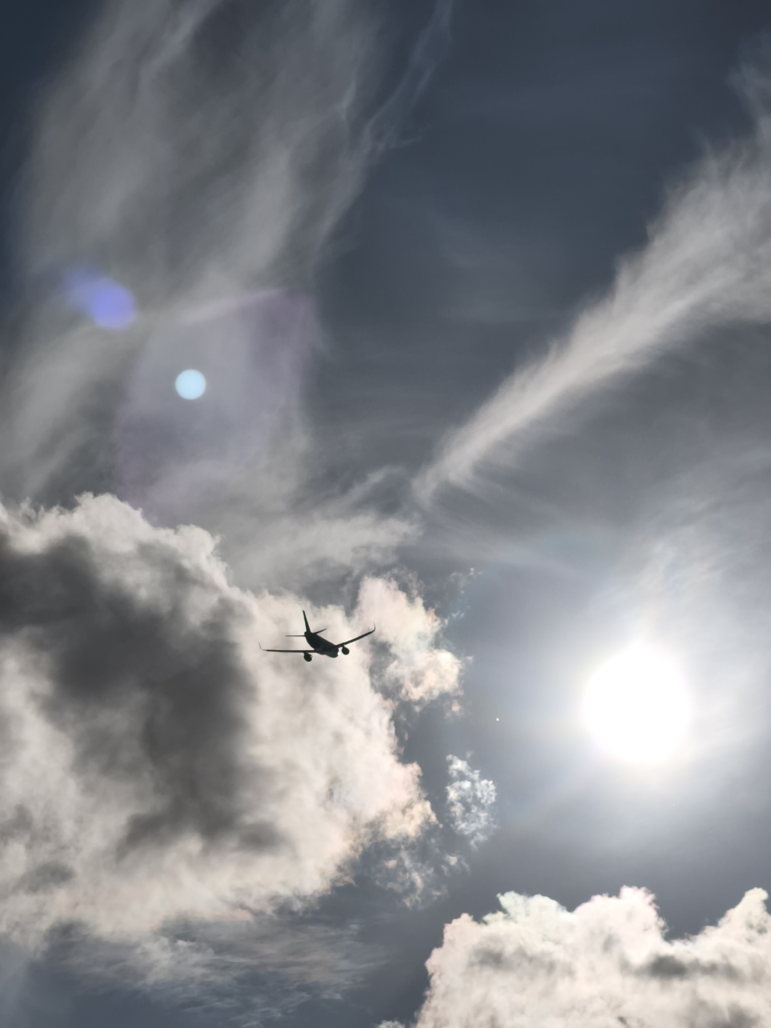 An airplane in silhouette is flying through a cloudy, overcast sky. The sun is visible and bright, creating lens flares and illuminating some of the clouds from behind. Wispy cirrus clouds are visible against the darker sky, with larger, more opaque cumulus clouds present lower down. The airplane is positioned in the lower center of the frame, appearing to fly towards the right. The scene captures a moment of air travel against a dramatic sky. There is no information in the image to suggest a specific location in Tallinn, Estonia.