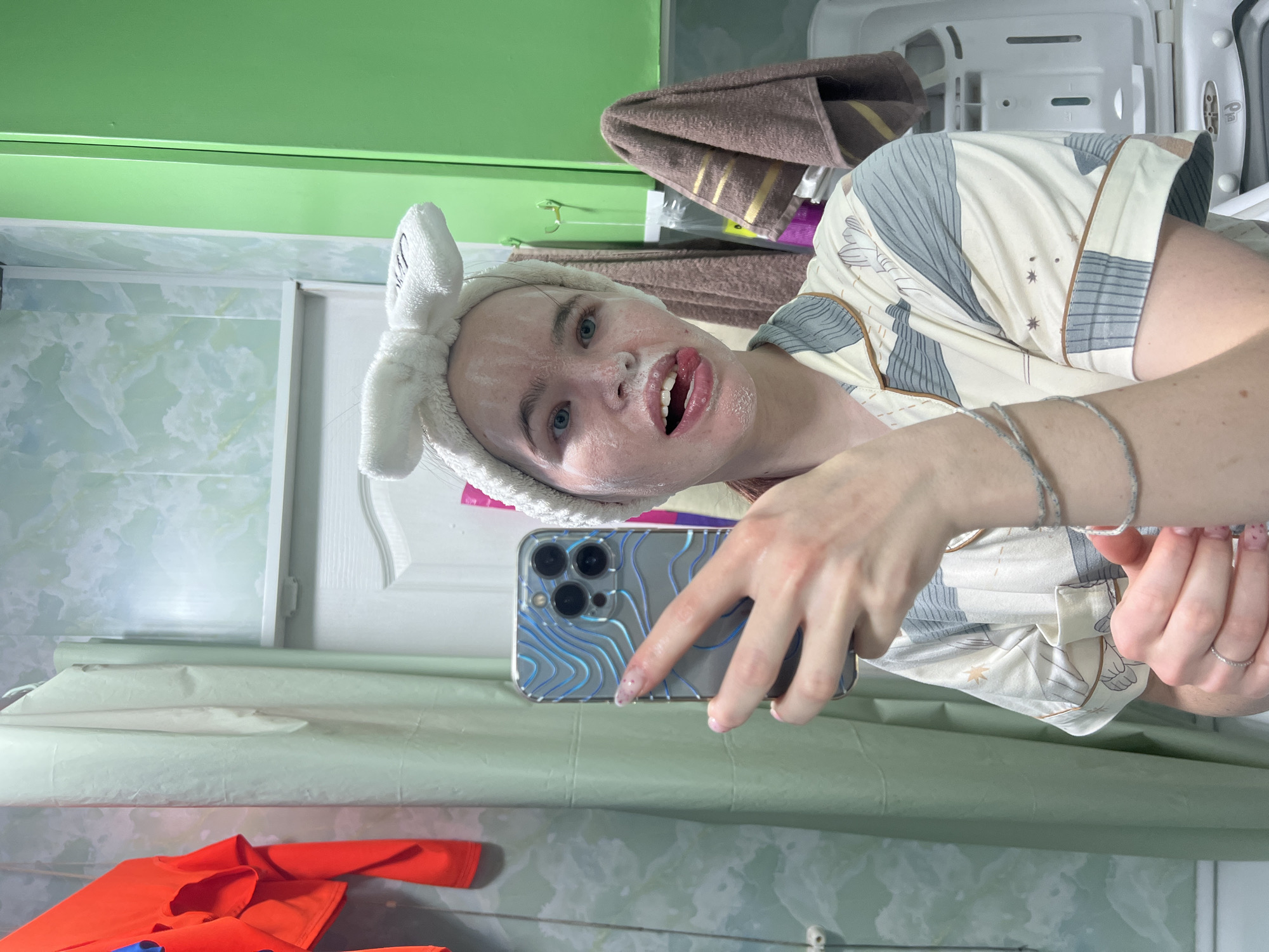 A selfie captures a young person, likely female, in a bathroom, engaged in a skincare routine. Their face is covered in a white, foamy substance, and they wear a white, fluffy bunny-ear headband. They have light skin, blue eyes, and a playful expression, with their mouth open and tongue slightly out.
The individual is wearing a cream-colored pajama top with a grey and brown geometric and abstract pattern. On their left wrist are multiple silver bracelets, and a simple ring adorns their left ring finger. They hold a smartphone, encased in a clear cover featuring blue wavy lines, in their right hand to take the photo.
The background reveals typical bathroom elements. A green painted surface or cabinet is visible at the top left, while the walls are adorned with light-colored tiles displaying a marbled, light green and white pattern. A white door is partially seen on the left. In the upper right, a brown towel with yellow stripes is draped, next to a white storage unit or open cabinet. Below the door, a roll of light-colored paper or fabric rests on a shelf, and an orange garment lies on a surface in the bottom left. The overall mood is candid and relaxed, capturing a moment of self-care. No visible text is present in the image.