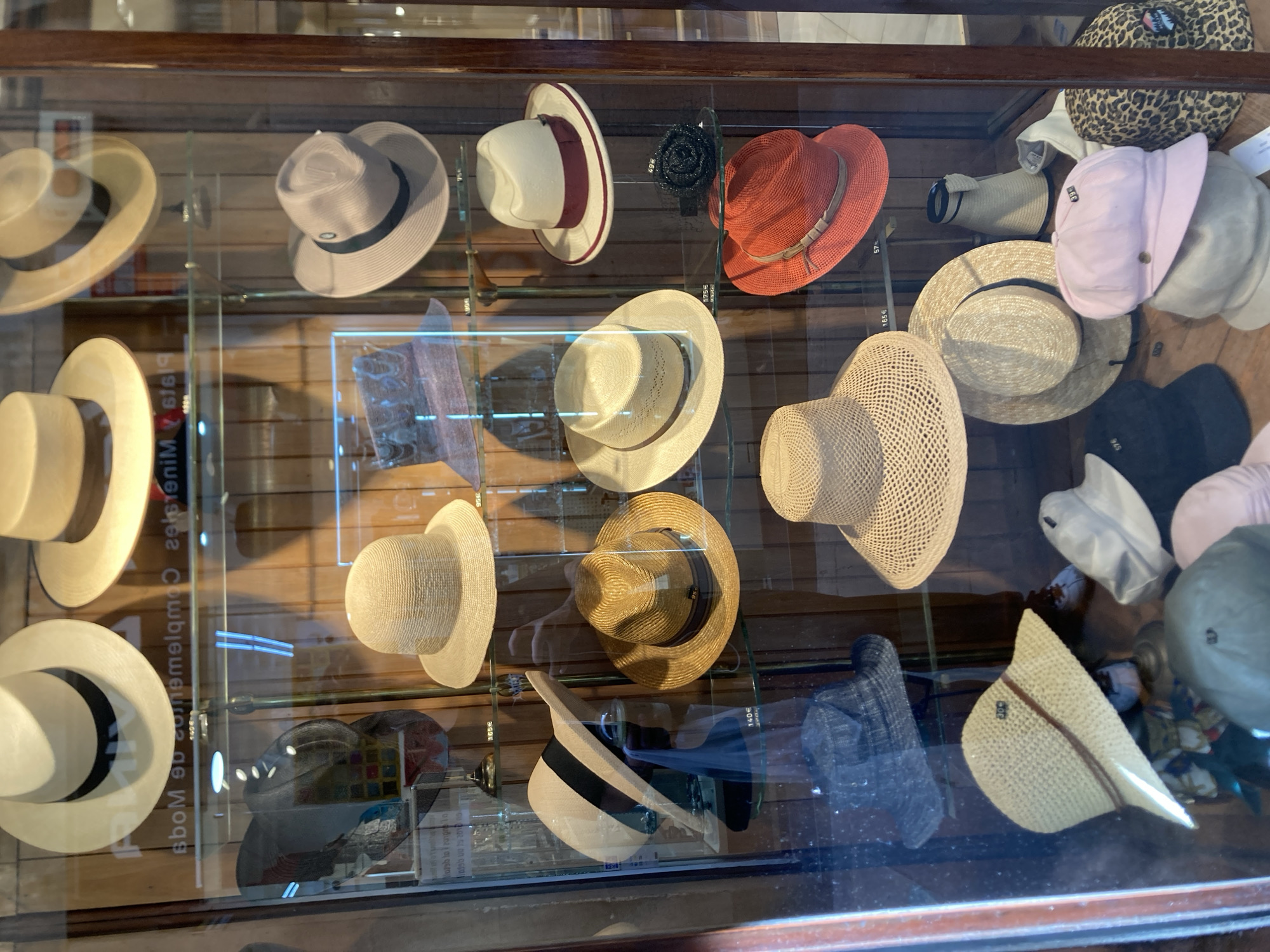 The image shows a display of various hats inside a glass-fronted cabinet, likely a shop window in Barcelona, Spain. The hats are arranged on shelves and are made from a variety of materials, including straw, woven fabrics, and possibly felt. Styles range from wide-brimmed sun hats to more structured fedoras. Some hats are plain, while others have decorative bands or ribbons.
The lighting suggests an indoor environment, possibly with natural light filtering through the glass. There are reflections on the glass, indicating it is a display viewed from the outside. Visible text on price tags or labels includes numbers and currency symbols like "€" and "65€," "140€," and "175€," suggesting these are items for sale. One label also shows "165€" and "57€". Another piece of text is visible, "Bbom e cometenim 61619," which is likely reflective from within the shop or a label. The setting appears to be a retail store specializing in hats. The overall impression is one of a well-organized and diverse hat collection on display.