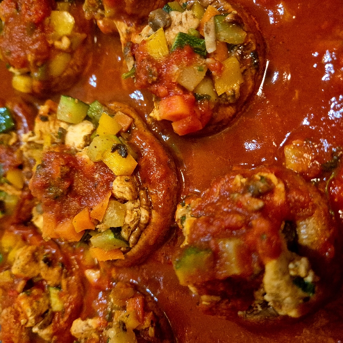 The image presents a close-up, high-angle view of multiple cooked stuffed mushroom caps arranged in a deep, rich red sauce. Approximately eight individual stuffed mushrooms are visible, partially submerged in the thick, viscous sauce, which appears to be tomato-based. Each mushroom cap is light to medium brown and filled with a colorful mixture of finely diced ingredients. Identifiable components within the stuffing include orange cubes (likely carrots or bell peppers), yellow cubes (likely bell peppers), and green pieces (likely zucchini, bell peppers, or herbs). Lighter, irregular pieces are also present within the stuffing, suggesting additional ingredients such as mushroom stems or cooked protein. Portions of the red sauce also rest on top of the stuffing in several mushrooms. The food is contained within an unseen cooking or serving vessel. The image provides no contextual information to determine its location within the city of Strasbourg, France. No people, objects other than food, or discernible actions are depicted.