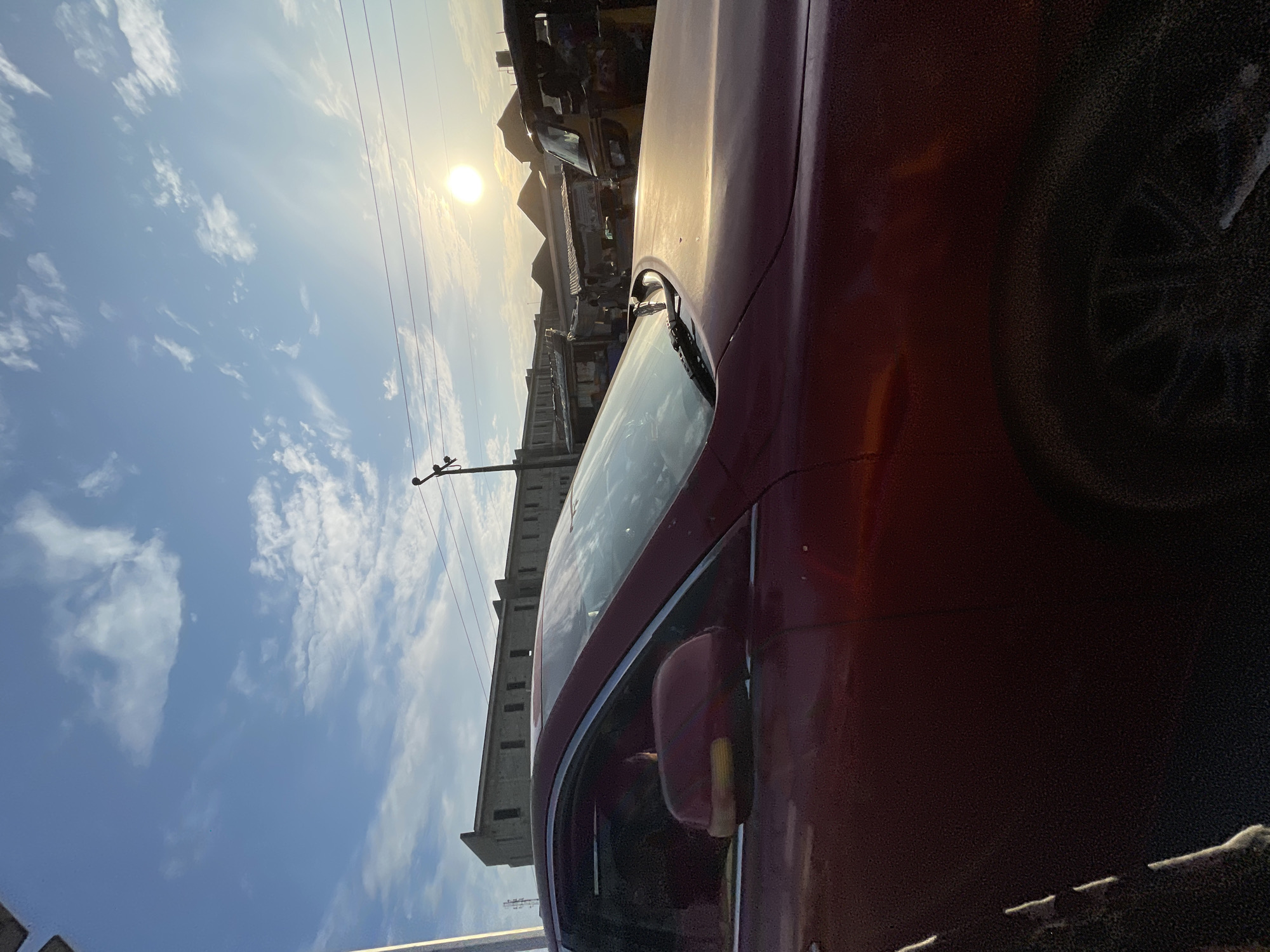 The image depicts a roadside scene in Ado, Nigeria, captured from the perspective of someone inside or beside a dark red car. The sun is bright and high in a partly cloudy blue sky, indicating it is daytime, likely midday. Power lines are visible stretching across the frame, and a utility pole with insulators is also present, suggesting an urban or semi-urban setting.
In the background, a long, multi-story building with many windows stands prominently. Across the street from this building, a row of smaller structures, possibly shops or stalls, are visible. A yellow auto-rickshaw, commonly known as a "keke napep" in Nigeria, is parked or moving in the distance, adding to the local context. The car in the foreground is dark red, and its side mirror is also visible. The overall scene is typical of a busy street in Nigeria, with vehicles, buildings, and infrastructure elements characteristic of the region. No specific activity or event is clearly discernible beyond the general presence of traffic and daily life. The lighting suggests a sunny day, and the atmosphere seems ordinary for the location.