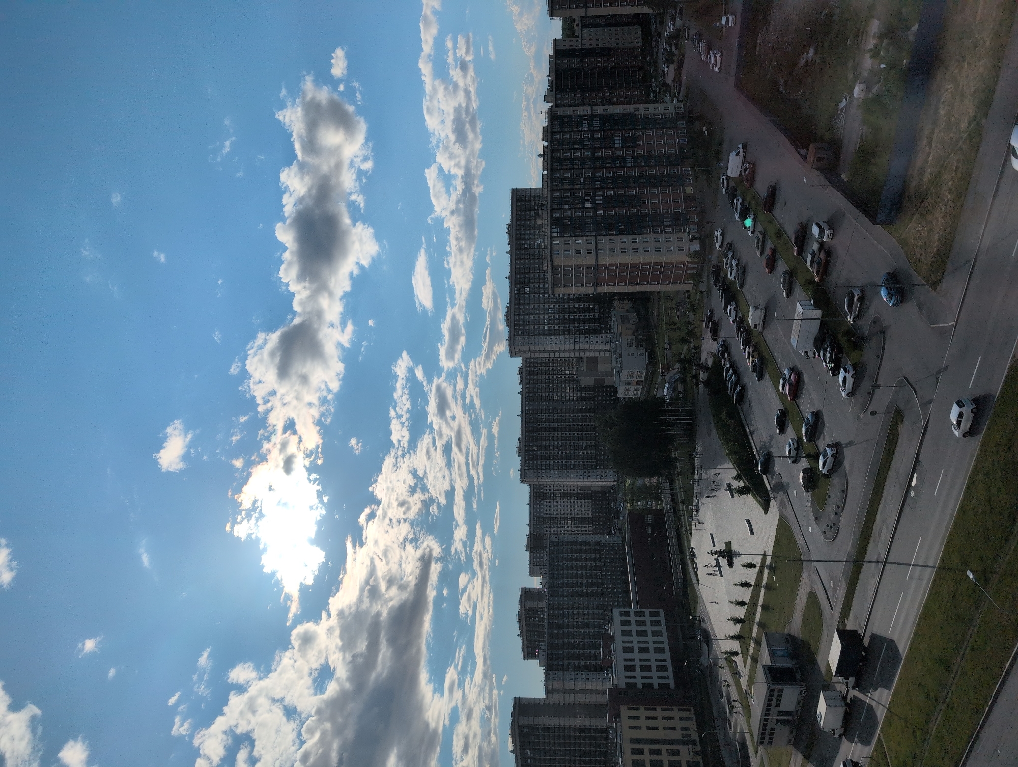This media file captures an aerial view of a residential area in Saint Petersburg, Russia, during what appears to be a sunny day with scattered clouds. The scene is dominated by numerous tall apartment buildings, characteristic of urban Russian architecture. Below the buildings, a road and parking lot are visible, with several cars parked and some in motion. The presence of a white van and a truck suggests ongoing activity such as deliveries or residential movement.
The time of day is likely afternoon, indicated by the bright sunlight and the angle of the shadows cast by the buildings and trees. The sky is a clear blue with fluffy white and gray clouds, suggesting fair weather with some cloud cover. The overall impression is one of a bustling, modern urban environment. There is no visible text in the image.