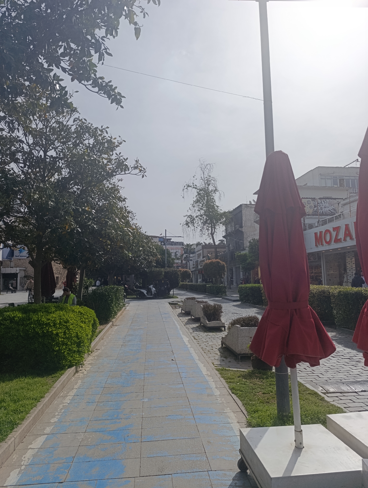 The photograph depicts a street scene in Antalya, Turkey. A paved walkway stretches into the distance, bordered by manicured hedges and trees on the left. On the right, there are closed red umbrellas, sidewalk planters, and buildings. The "MOZA" store is visible in the background. Several people are visible, including individuals walking and sitting along the street. Motorbikes are parked along the side. The sky is overcast, indicating daytime.
