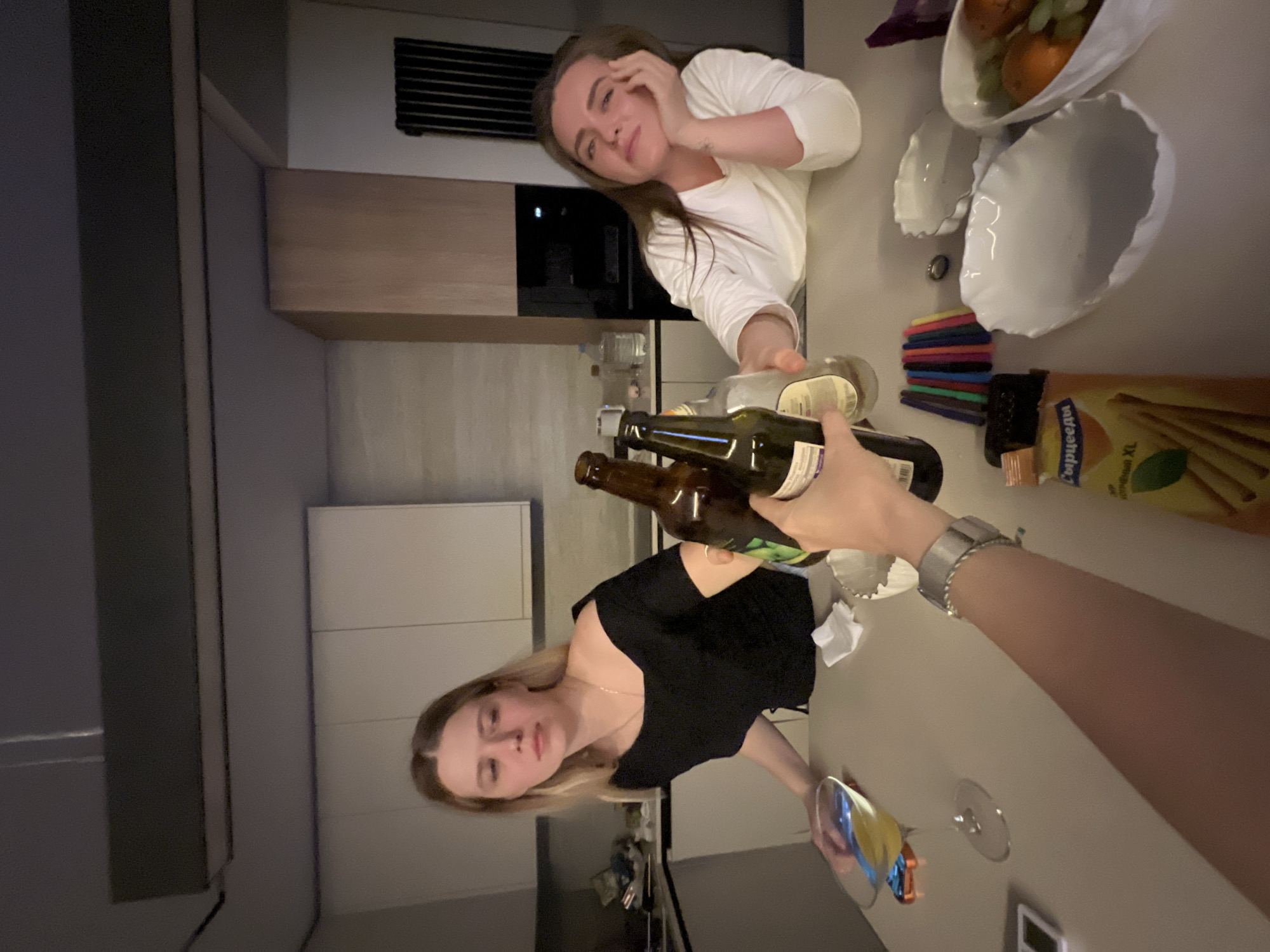 The image captures a scene in what appears to be a modern kitchen. Two women are visible. One, with blonde hair and wearing a black top, is positioned lower in the frame, holding a clear wine glass. The other woman, with brown hair and wearing a white top, is positioned higher in the frame with her head resting on her hand.
Various objects are scattered on the countertop: a brown beer bottle, a green wine bottle, a half empty bottle of white wine, colored pens, a bag of snack sticks, and a fruit bowl. A silver watch is visible on an arm holding the wine bottles.
The kitchen features light-colored cabinets and countertops. A microwave and air vent are visible. The overall ambiance suggests an informal gathering. There is no evidence to definitively link the scene to the city of Рубцово, Russia.