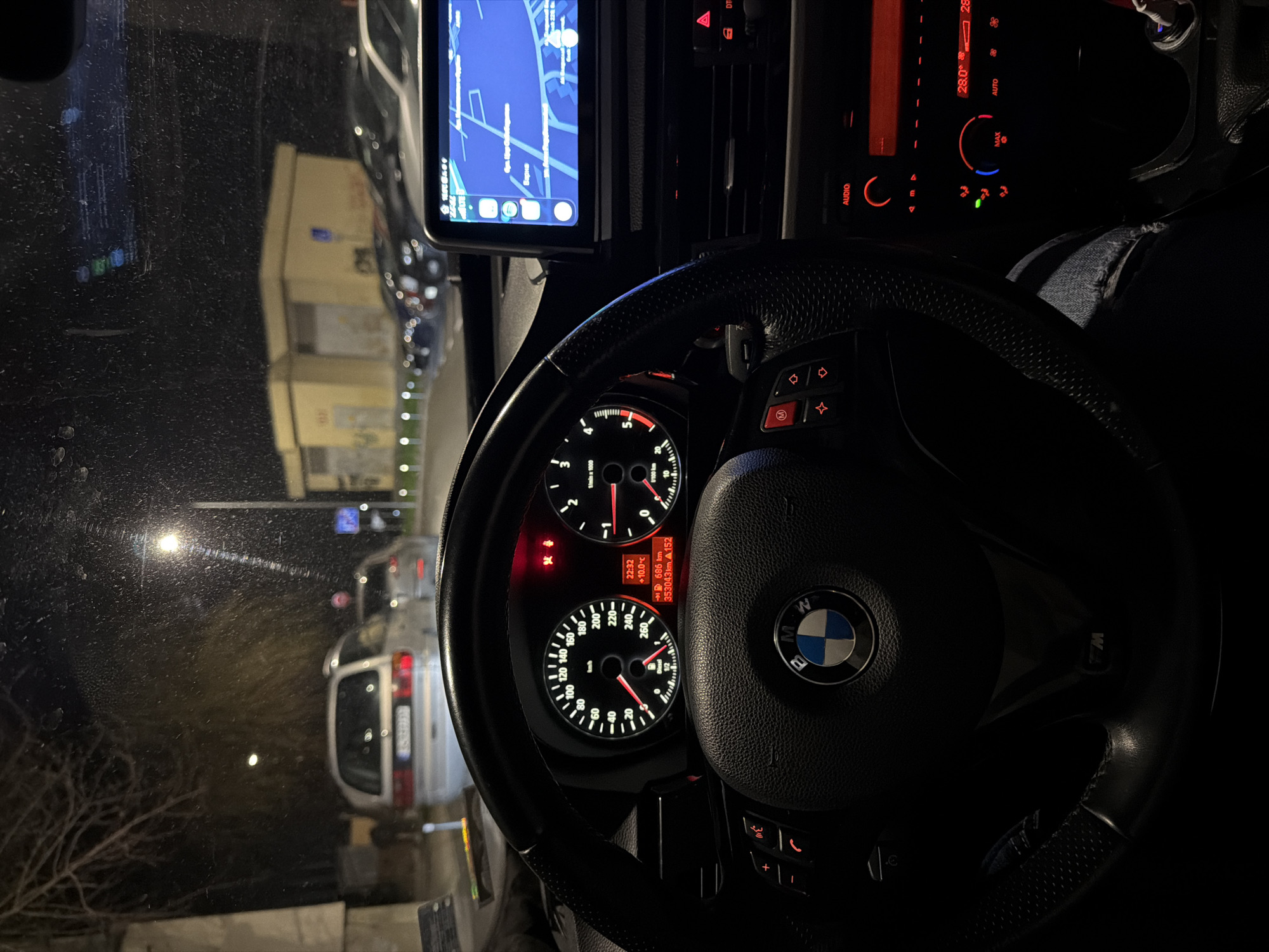 The image depicts a nighttime scene from the interior of a BMW vehicle, looking outwards through a dirty or speckled windshield. The steering wheel, displaying the BMW logo and an "M" logo, is prominently centered. The instrument cluster is illuminated, showing a speedometer with a needle at 0 km/h, a tachometer, and a digital display indicating the time as 22:32, an outside temperature of +10.0°C, a remaining range of 686 km, and an odometer reading of 353043 km. Red warning lights for a battery and possibly an airbag or seatbelt are active. To the right, an infotainment screen displays a navigation map with "Варна" (Varna) clearly visible in Cyrillic text, confirming the city in Bulgaria. Below the screen, the climate control panel is illuminated in red, showing a temperature setting of 28.0°C. A portion of the driver's leg, clad in blue ripped jeans, is visible in the lower right foreground.
Outside the vehicle, a light-colored station wagon or minivan is parked or idling directly ahead, with its rear lights visible. Additional vehicles are discernible further down the street. In the middle distance to the right, a distinctive light yellow, rectangular building with minimal fenestration is visible. The scene is lit by streetlights, one of which is prominent directly ahead and slightly right. Bare tree branches are visible against the dark sky on the left. The overall environment appears to be an urban street or parking area in Varna, Bulgaria.