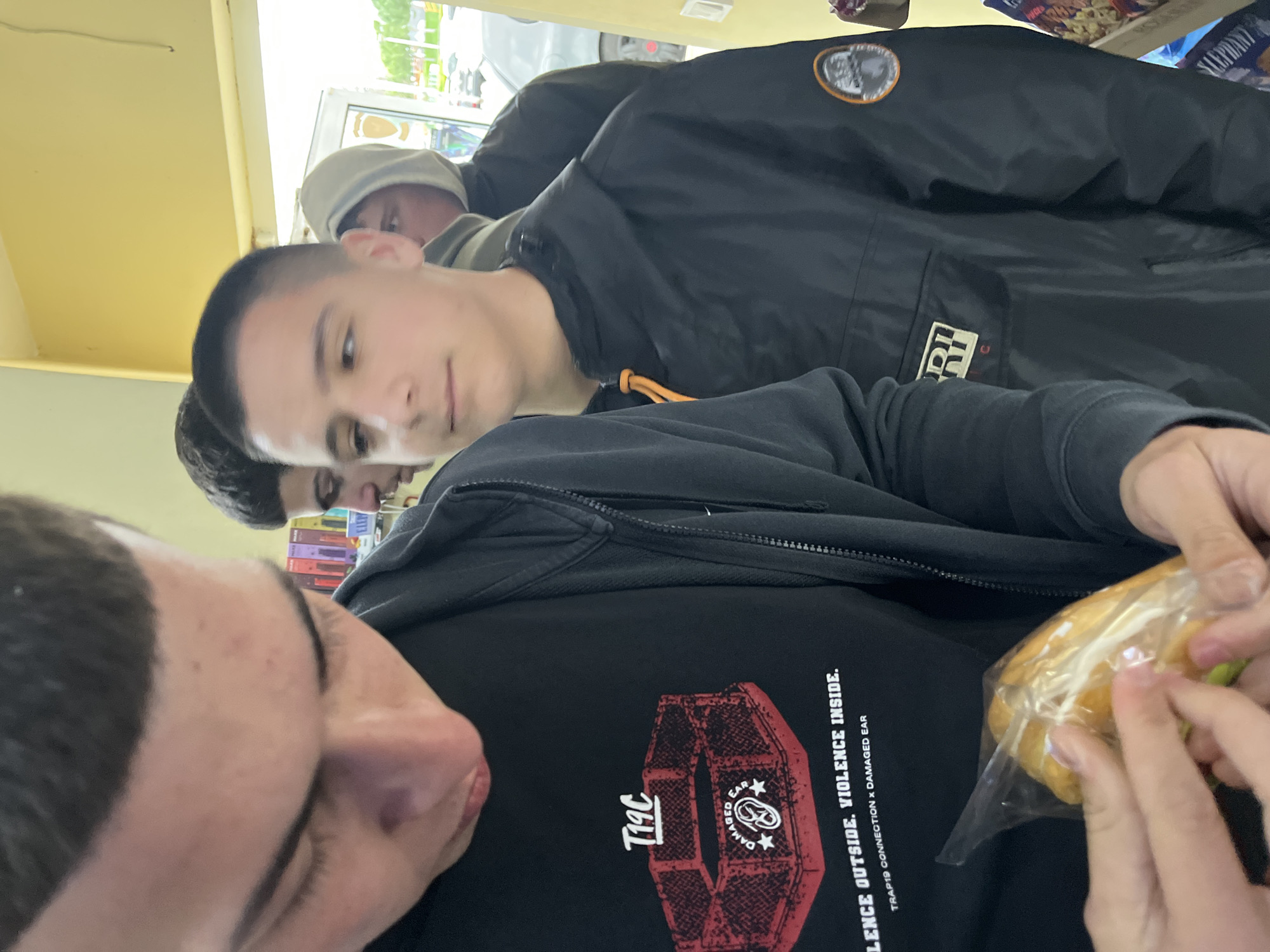 The image shows a group of young men indoors, possibly in a store given the visible window and shelf contents. The individual closest to the camera is wearing a black t-shirt with a graphic design featuring the text "T19C" and "VIOLENCE OUTSIDE. VIOLENCE INSIDE." He is holding a wrapped sandwich. Two other young men are visible behind him, with one wearing a grey hat and another wearing a black jacket with a patch. The environment is generally light, with a yellowish tint from the indoor lighting. The location in Burgas, Bulgaria, cannot be determined from the visual information provided.