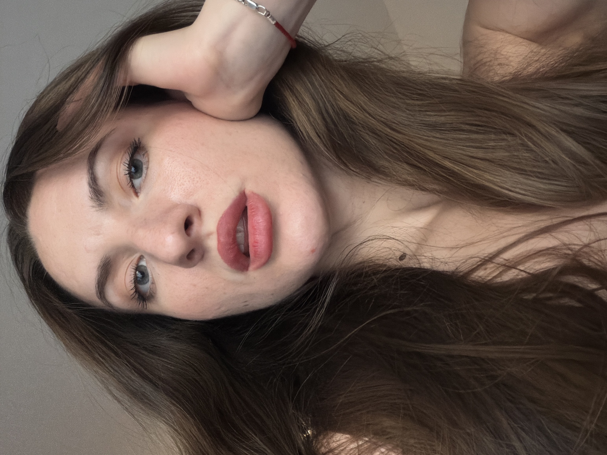 The image is a close-up, high-angle, selfie-style photograph of a young woman's face and upper body. She has long, brown hair that is partially obscuring her face and shoulder. Her left hand is resting on her cheek, and she is wearing a thin red bracelet with silver beads on her wrist. Her lips are painted a coral-pink shade, and her eyes are looking upwards and slightly to the right. Her skin appears fair with some freckles visible on her cheeks. The background is a plain, light-colored wall. The photograph does not contain any elements that would suggest a location in Sharm El Sheikh, Egypt.