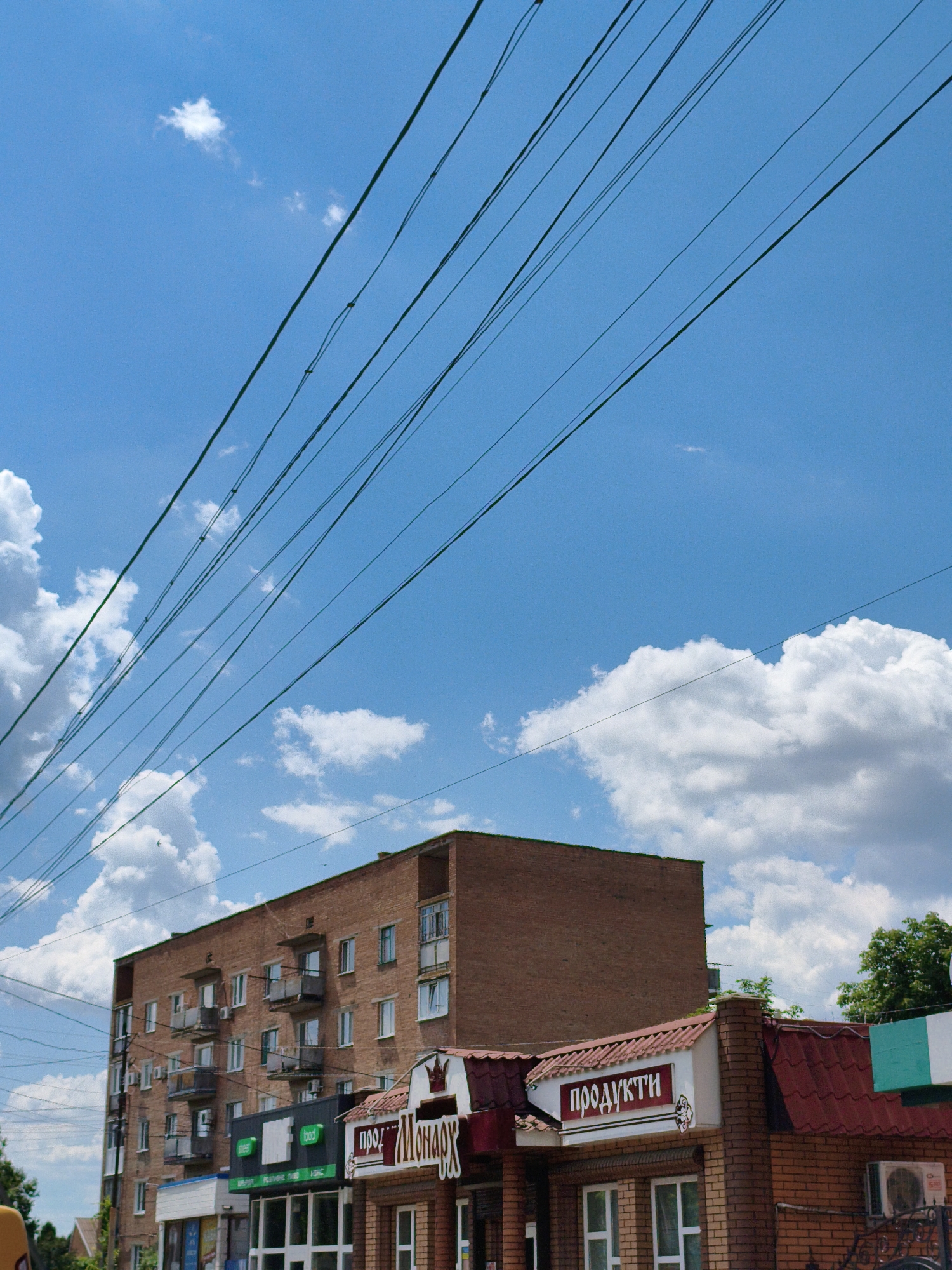 This image captures a street view in Velyka Bahachka, Ukraine, on a sunny day. A multi-story brick apartment building dominates the background, with several floors featuring small balconies. Below the apartment building, a row of storefronts is visible, including one with a sign that reads "ПРОДУКТИ," indicating a food store. Power lines stretch diagonally across the bright blue sky, which is dotted with fluffy white clouds. The scene suggests a typical urban environment in a Ukrainian town, with a mix of residential and commercial structures.