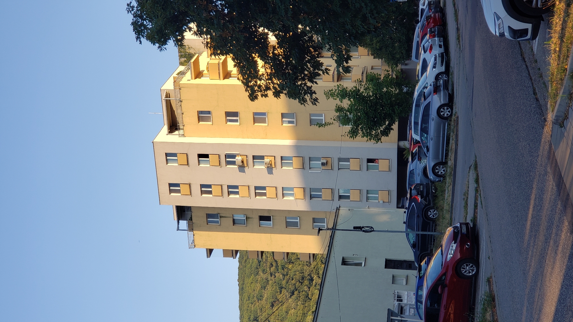 A tall, multi-story apartment building with a yellow and grey facade stands against a clear blue sky. The building has several balconies and numerous windows. In front of the building, a parking lot is filled with various cars, including a red hatchback, a black SUV, and several silver sedans. To the left of the apartment building, a shorter, light-colored building is visible. The background features a lush green, tree-covered hillside. The scene appears to be an urban residential area, likely a neighborhood in Bratislava, Slovakia, given the license plate on the red car, which reads "BL 177LX". The sunlight casts long shadows, indicating it is either early morning or late afternoon. One person is visible on a balcony on the lower floors of the apartment building.