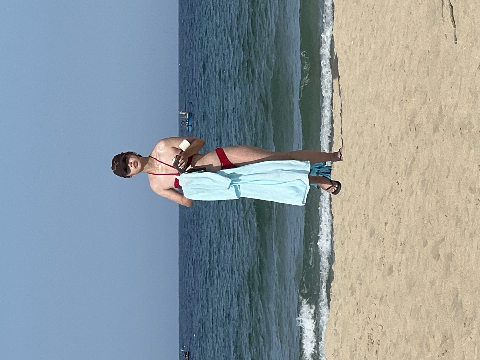 A young woman stands on a sandy beach in Đà Nẵng, Vietnam, facing the camera. She is wearing a red bikini and a dark baseball cap. She holds a light blue towel draped over her left arm and a small brown object, possibly a phone or a wallet, in her right hand. Her skin appears slightly sun-kissed. The ocean stretches out behind her, with gentle waves lapping at the shore and a few small boats visible in the distance. The sky above is a clear, bright blue. The sand is light brown and appears dry, with subtle footprints.