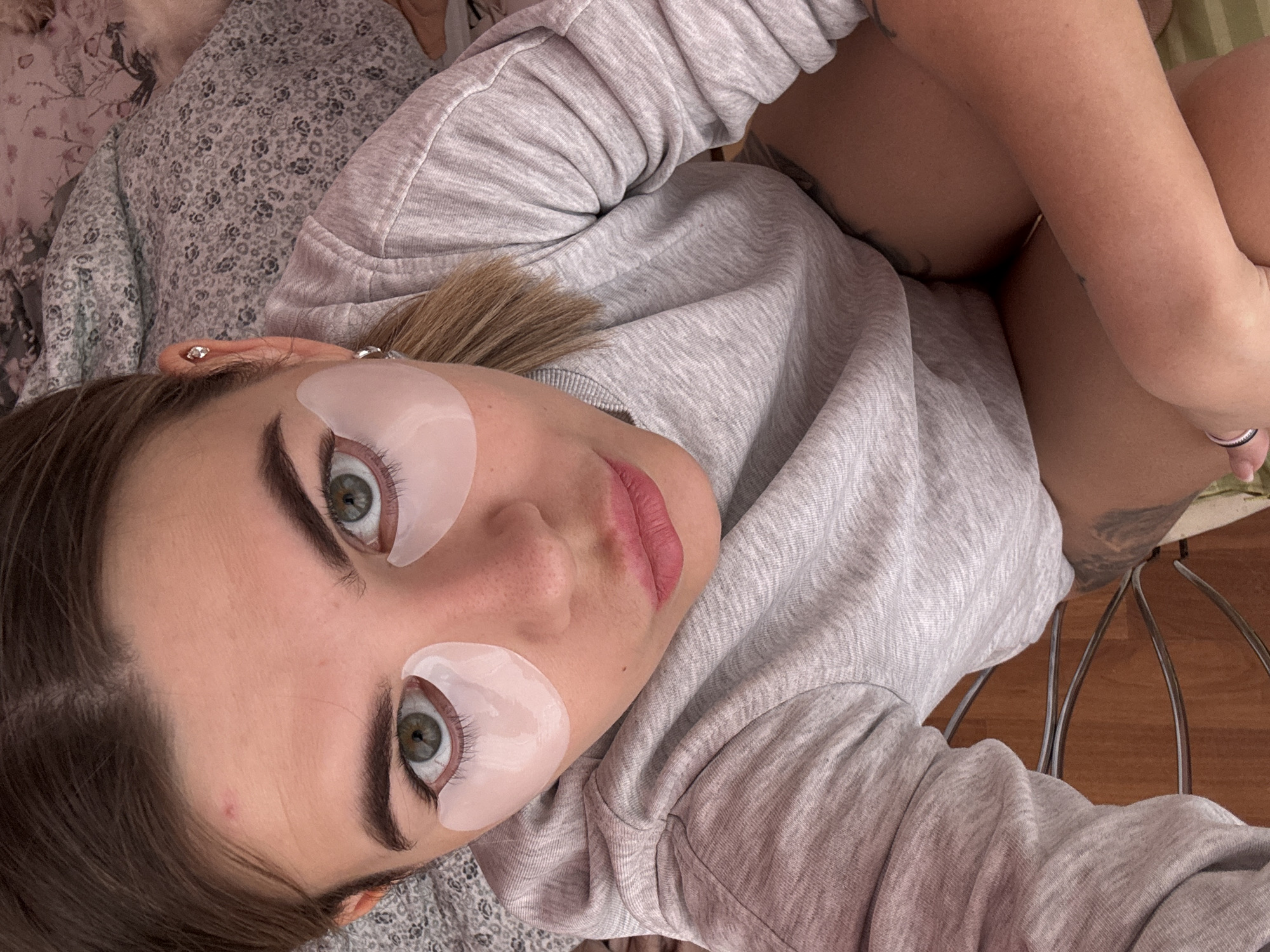 The image depicts a young adult female indoors, looking directly into the camera from an elevated angle. The specific location within Chaikovsky, Russia, cannot be determined from the visual information provided. She has fair skin, brown hair parted down the middle, and green eyes. Two white, translucent hydrogel patches are applied beneath her lower eyelids. Her eyebrows are dark and defined, and her lips are painted with a matte pink-mauve lipstick. She is wearing a light grey, long-sleeved crew-neck sweatshirt. A silver stud earring is visible in the helix of her left ear, and a small silver hoop earring is in her left earlobe. A dark tattoo is partially visible on her upper right thigh. She appears to be reclining against patterned bedding with floral and botanical motifs, visible in the upper right quadrant of the image. Below her, partially visible, is a piece of furniture with a curved, metallic-looking backrest, possibly a chair. Her right arm is bent, with a ring visible on one finger. The environment suggests a domestic setting, likely a bedroom or living space.