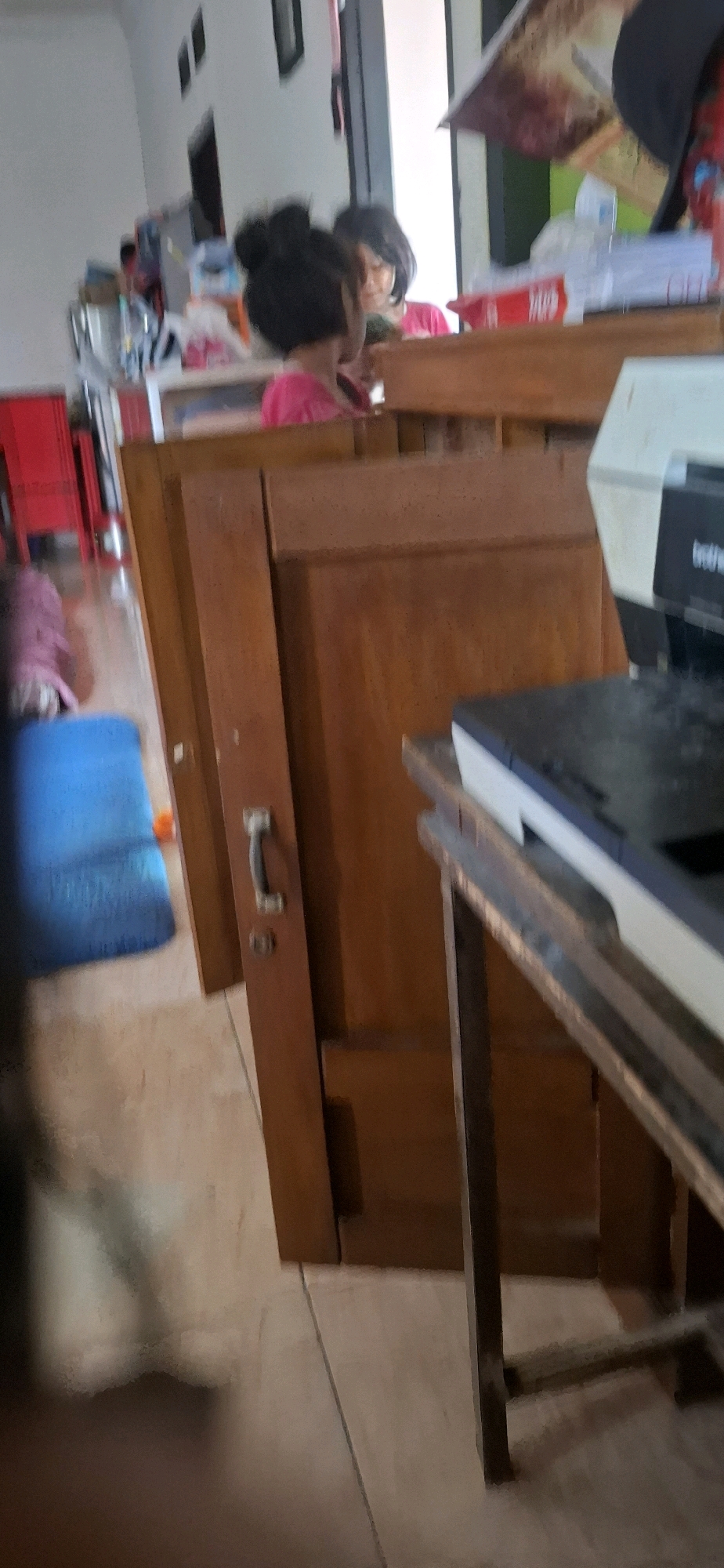 The image shows an indoor scene, likely a residential setting in Cibinong Hilir, Indonesia. In the foreground, a wooden door with a dark handle is visible. To the right, a printer with "brother" branding is placed on a table with metal legs. A blue mat is on the floor to the left. In the background, a young girl with her hair in buns and wearing a pink shirt is reflected in a mirror, looking towards the right. Another person, partially obscured, is also in the background. The environment appears to be a room with white walls and tiled flooring.