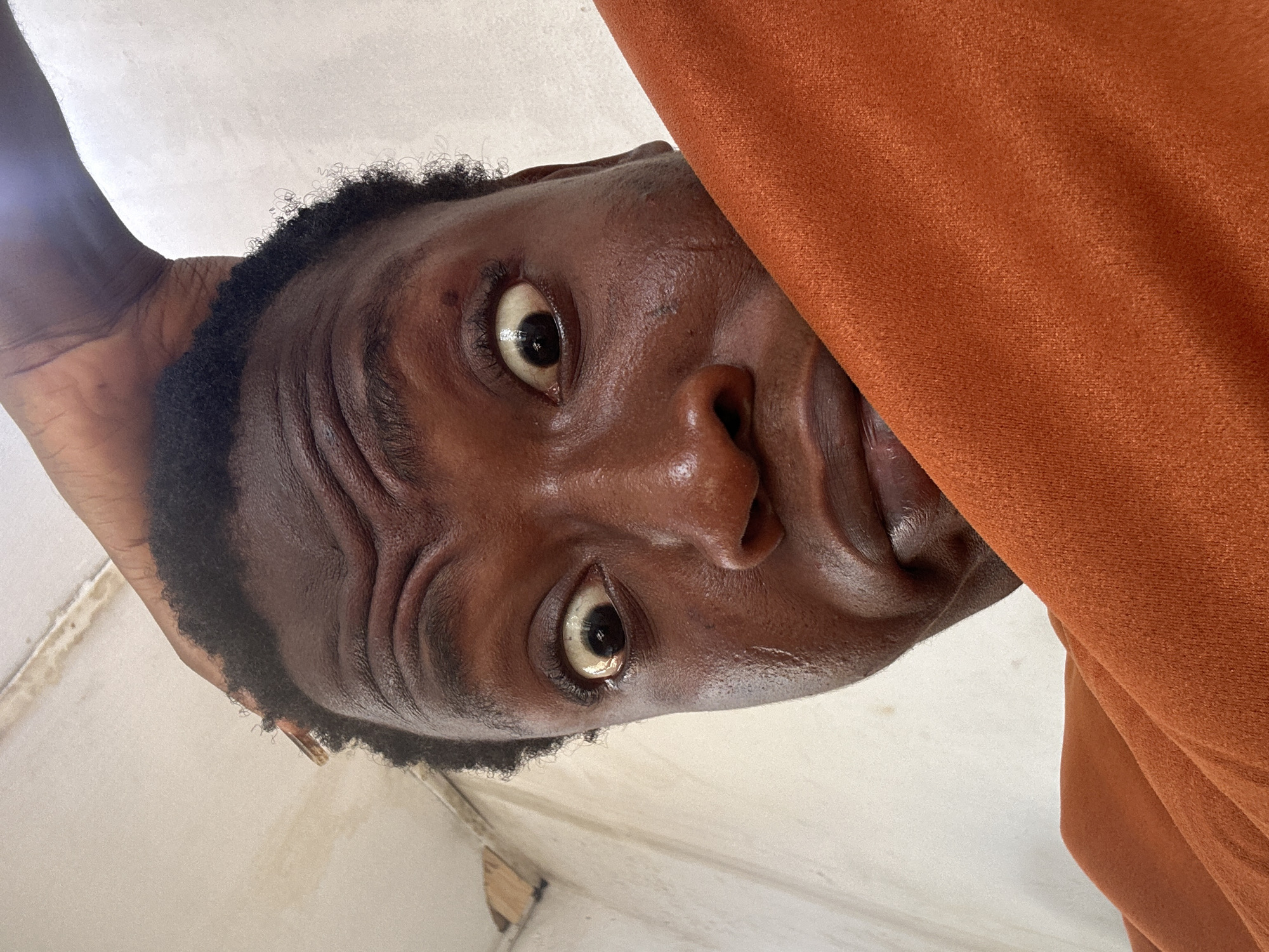 A low-angle close-up photograph shows a person with dark brown skin looking down at the camera. The person has short, dark hair and prominent eyebrows. Their eyes are wide open, and the expression on their face appears curious or surprised. Part of an orange fabric is visible on the right side of the frame, obscuring some of the person's face. The background appears to be a plain, light-colored wall. The location is in the city of Unknown, Unknown.