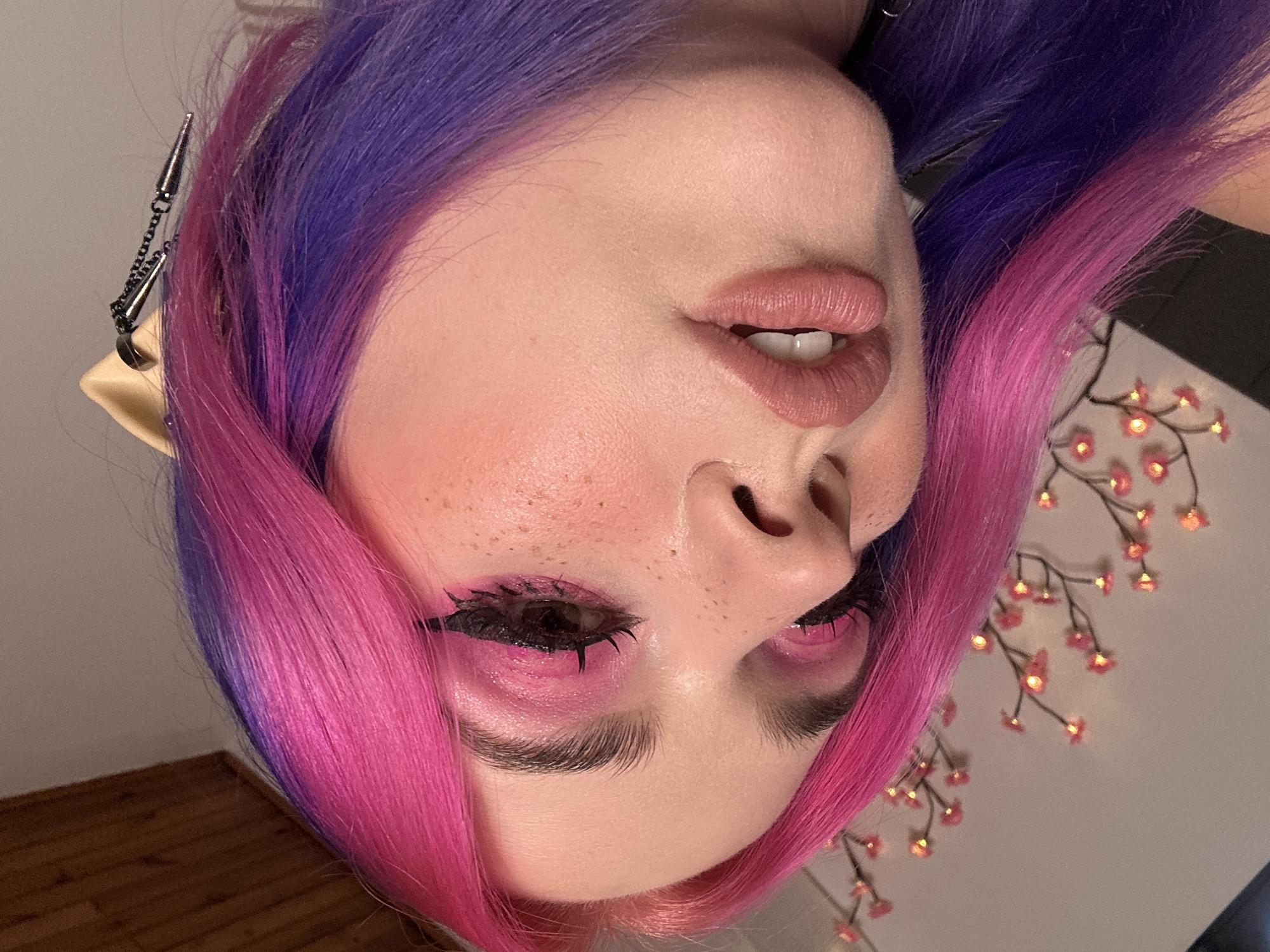 A close-up shot of a person's face. The person has vibrant, multi-colored hair, transitioning from purple at the roots to pink at the tips. They have light skin with pinkish tones on their cheeks, possibly blush. Their eye makeup is also pink, with defined black eyeliner and lashes. The person appears to have elf ears.
There are some floral string lights in the background. The flooring is made of wood. No information about the location in the city of Zagreb, Croatia, can be inferred from this photo.