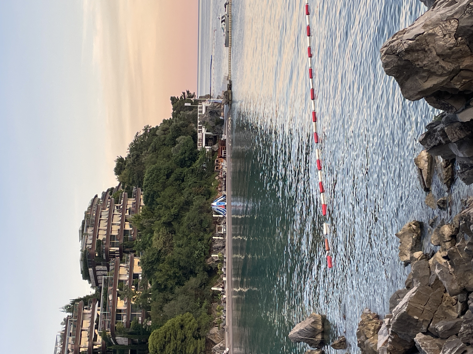 The image captures a tranquil scene at a beach resort in Budva, Montenegro, likely during the early morning or late evening, as indicated by the soft, warm hues of the sky. In the background, a multi-tiered hotel complex with modern architecture is nestled into a lush, green hillside. The hotel features numerous balconies and terraces adorned with vegetation, blending seamlessly with the natural landscape.
In the middle ground, a pebble beach is visible, furnished with lounge chairs, umbrellas, and a small, tent-like structure. The calm, turquoise waters of the Adriatic Sea gently lap the shore. A rope barrier with red and white buoys delineates a swimming area in the foreground. The water is remarkably clear, with large rocks protruding from it near the shoreline, adding to the natural beauty of the setting. A small boat is moored to a floating dock in the distance, suggesting a marina or a dedicated spot for water activities. The overall impression is one of serene beauty and relaxation, characteristic of the Montenegrin coast.