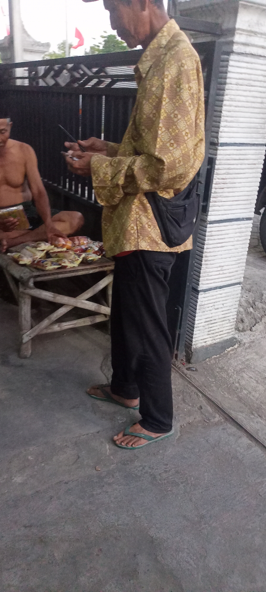 In Gempolklutuk, Indonesia, a man in a gold patterned long-sleeved shirt and black pants, wearing green flip-flops, stands on a concrete surface. He is holding a mobile phone in his right hand and appears to be interacting with it. To his left, another man, shirtless, sits on a wooden stool next to a table displaying an assortment of packaged goods. The table is made of light-colored wood and has a criss-cross leg design. Behind the men is a black metal fence, and beyond that, a white striped wall. Red flags are visible in the background.