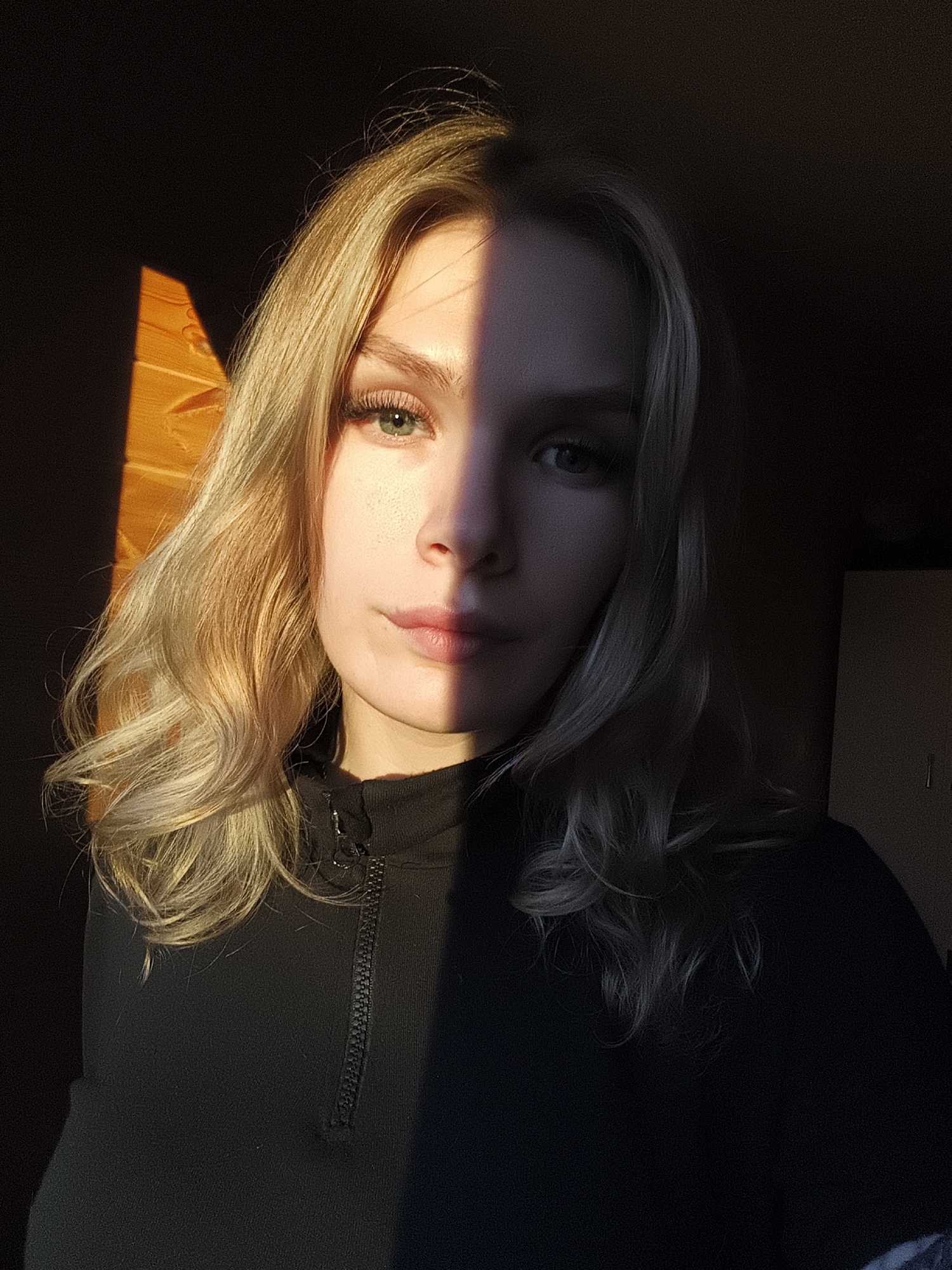 A striking close-up portrait features a young woman with blonde, wavy hair, dramatically illuminated indoors by a warm sunbeam. Her face is split by a sharp line of light and shadow, with her left side (viewer's right) bathed in golden sunlight, revealing green eyes, long eyelashes, and subtle freckles on her cheek and nose. Her right side (viewer's left) is in deep shadow. She maintains a direct, neutral gaze towards the camera.
She is wearing a black, high-necked top with a visible half-zip detail. The strong, low-angle sunlight suggests it is either late afternoon or early morning. In the background, on the sunlit left side, textured wooden slats are visible. On the dimly lit right side, an indistinct light-colored cabinet or piece of furniture can be faintly discerned in the darkness. There is no visible text in the image. The setting is an indoor environment in Русский Вожой, Russia.
