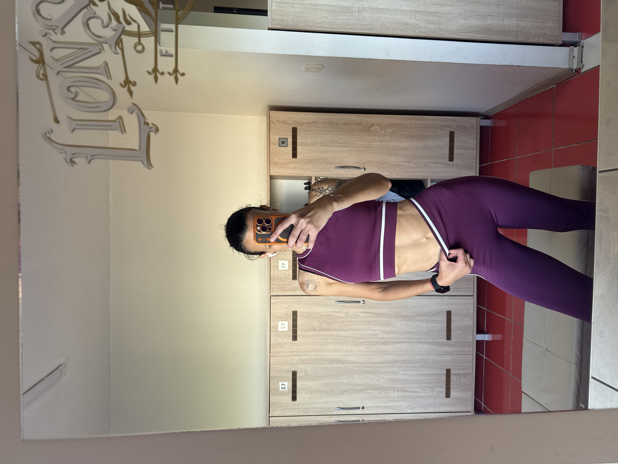 The image depicts a woman taking a mirror selfie in a changing room. She is wearing a purple sports bra and leggings with white stripes. The changing room has lockers with numbers on them. The floor is tiled in red and beige. A decorative sign hangs in the upper left corner of the mirror, partially obscured by the frame. The location appears to be in Pleven, Bulgaria, based on visual cues such as architectural style and signage.