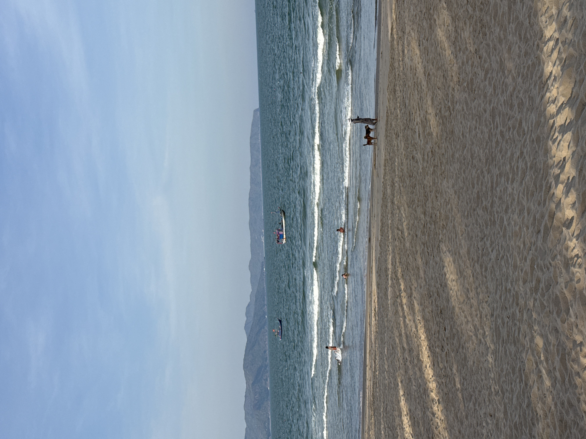 This is a landscape-oriented photograph of a beach scene in South Quy Nhon Ward, Vietnam. The image captures a wide expanse of sandy beach meeting the ocean, with mountains visible in the distance. The sun is casting long shadows across the sand, indicating it is likely late afternoon.
Several people are present in the water and on the beach. Two dogs are standing on the wet sand near the water's edge, with one appearing to be looking out at the sea. A small boat with people on board is further out in the water, and another, smaller boat with a figure visible is closer to shore. Other individuals can be seen wading in the shallow waves. The water is a calm blue-green, with gentle waves rolling in.
The sky is a pale blue with faint, wispy clouds, suggesting a clear and pleasant day. The overall atmosphere appears relaxed and leisurely, consistent with a typical beach outing. There is no visible text in the image.