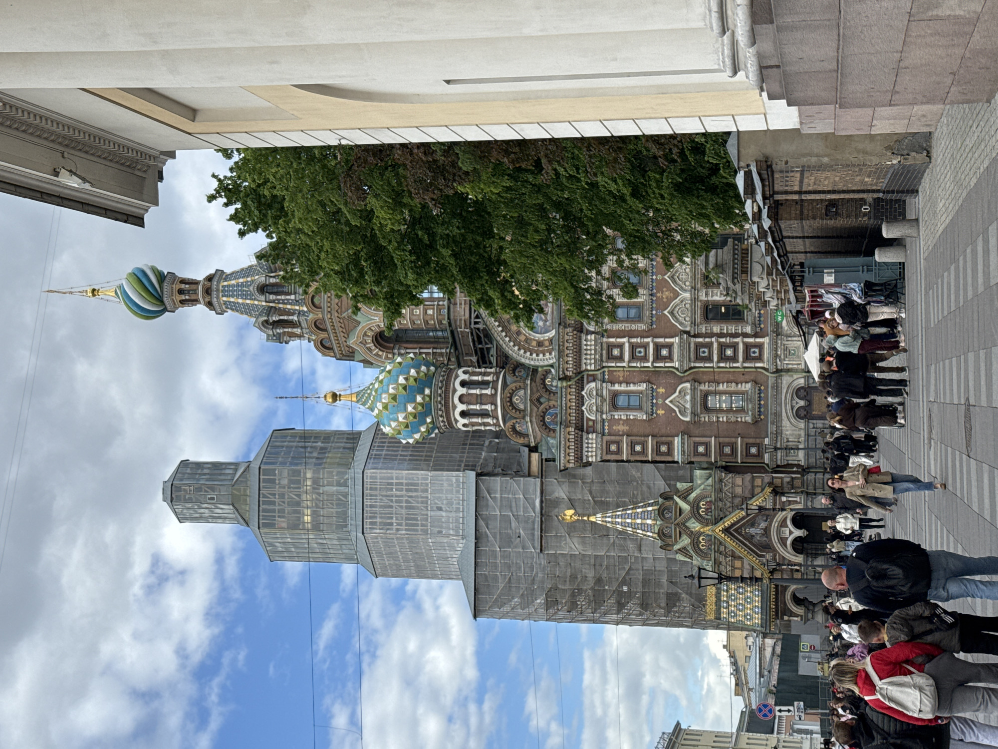 The image depicts the Church of the Savior on Spilled Blood in Saint Petersburg, Russia. Scaffolding obscures a portion of the building. A crowd of people is on the sidewalk. The sky is cloudy.
