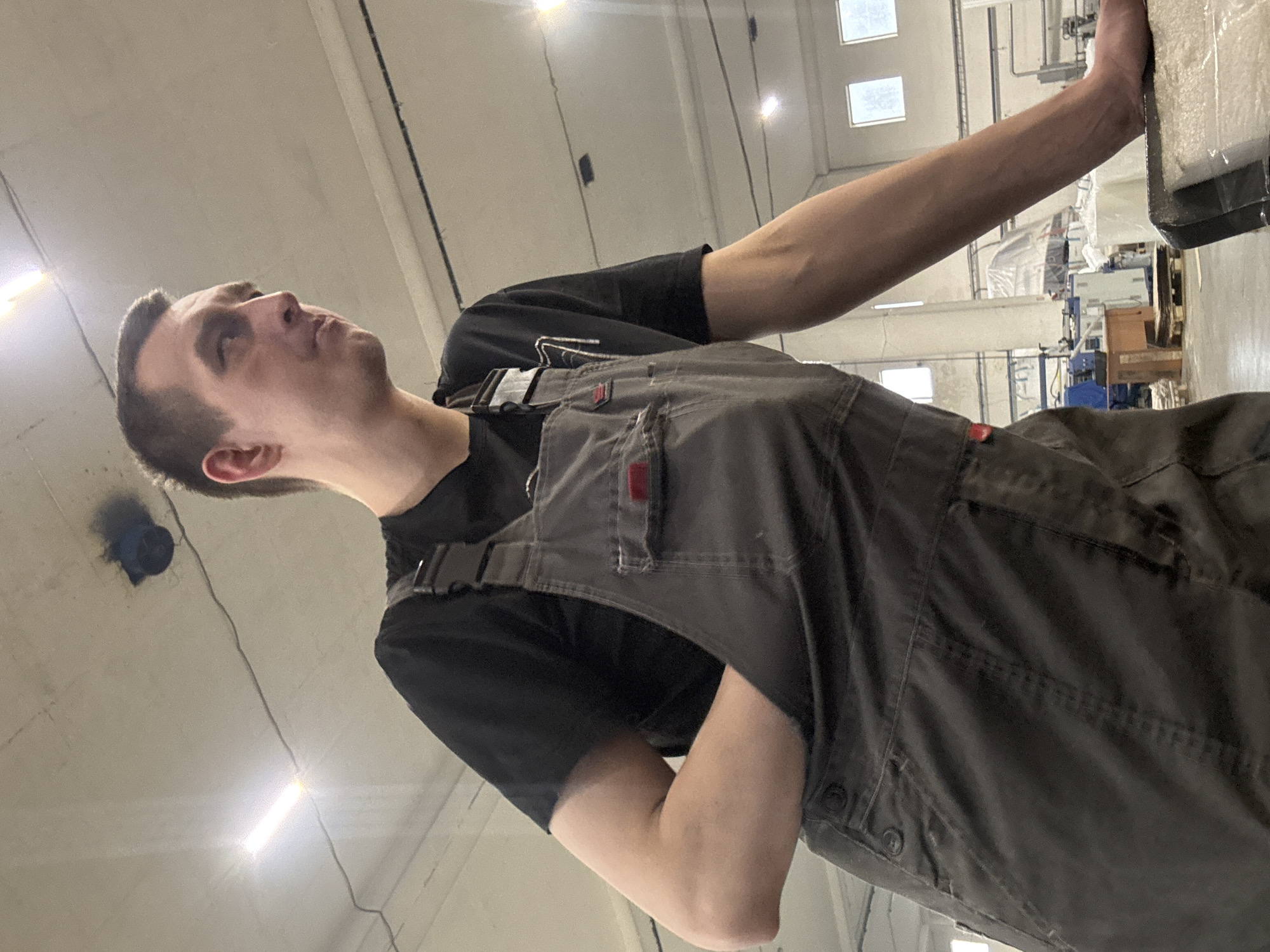 An adult male stands in an industrial facility in Dikaya Gar', Russia. He is positioned in a three-quarter profile, looking towards the right side of the frame with a contemplative expression. He wears a black t-shirt beneath dark grey work overalls featuring chest pockets, buckles on the shoulder straps, and buttons on the side. His right arm (viewer's left) rests flat on a textured, light-colored surface in the foreground, possibly a workbench or stacked material covered in plastic. His left arm (viewer's right) exhibits a significant limb difference, with the forearm appearing truncated or folded into the side pocket of his overalls, terminating above the elbow. The background reveals a high-ceilinged room with several rectangular fluorescent light fixtures, some brightly illuminated, and overhead electrical wiring. A circular dark blue fixture with an associated dark stain is visible on the ceiling. Further back, through rectangular windows, external light is apparent. Various industrial objects, including machinery covered in white plastic sheeting, a wooden pallet, and what appears to be a small wooden cabinet, are discernible in the deeper background. The facility walls are light-colored.