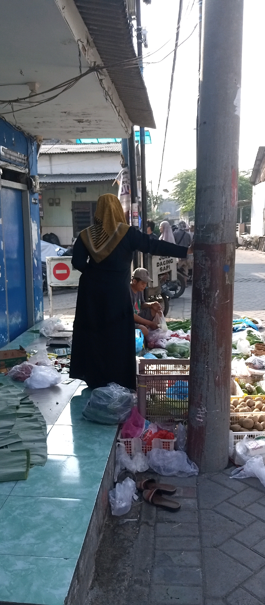 The image depicts an outdoor market scene, likely on a street, with a focus on a stall selling produce. A woman wearing a yellow hijab and a dark abaya stands near the center, her back mostly to the camera, with her right arm extended towards the right of the frame, possibly interacting with someone or something out of view. In front of her, on the tiled floor, are various items wrapped in plastic bags, including what appears to be produce. A metal scale is visible behind her.
To the right of the woman, a person wearing a baseball cap is seated, partially obscured by the woman's abaya, and is holding a plastic bag. Adjacent to them is a white motorized vehicle with text that reads "JUAL DAGING SAPI" (selling beef). Further to the right, a utility pole is prominent. The ground is paved with interlocking bricks, and more produce is displayed in crates and plastic bags, including potatoes and other vegetables. In the background, several other people are visible, some wearing hijabs. Buildings with corrugated metal roofs are present in the background. The scene suggests a commercial area, likely a street market, with evidence of commerce in fresh produce and meat. The location is identified as Wadungasri, Indonesia, indicating a tropical or subtropical environment, suggested by the lighting and the type of produce.