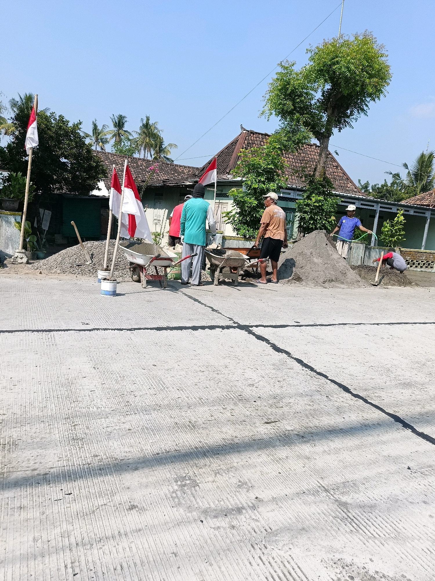 The image depicts a street construction scene in Manisrenggo, Indonesia, likely in front of a residential area. The foreground shows a freshly poured concrete road with a grooved texture. In the middle ground, several individuals are engaged in construction work. Two Indonesian flags are prominently displayed on bamboo poles, suggesting a national celebration or event. One flag is a larger national flag, while two smaller ones are also visible. Construction materials, including piles of gravel and sand, are present. Two wheelbarrows, one containing cement, are positioned near the materials. Workers are seen using shovels and a hose, indicating active construction. The background features traditional Indonesian houses with tiled roofs and lush greenery, including palm trees, under a clear blue sky. The overall atmosphere suggests a community effort towards infrastructure development.