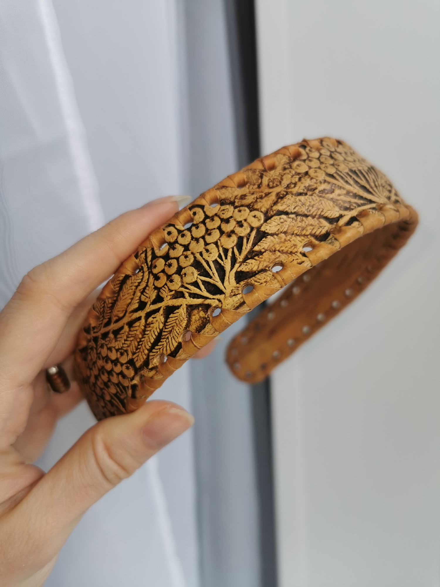 A person's hand holds a wide, intricately decorated headband or cuff, likely made from birch bark, which is light tan in color. The object is adorned with elaborate patterns of dark brown or black, created through pyrography or carving, depicting clusters of berries and leafy branches. Small, rounded cut-out holes are visible along the edges of the accessory. The interior surface is a plainer light brown with a series of small, evenly spaced holes, possibly for adjustment or a lining. The hand has fair skin and long, manicured fingernails, with a simple metallic ring on one finger. The background is softly blurred, showing a translucent white curtain or fabric on the left, suggesting an indoor setting with natural light, and a plain white surface on the right. The image focuses on the detailed craftsmanship of the item. No text is visible.