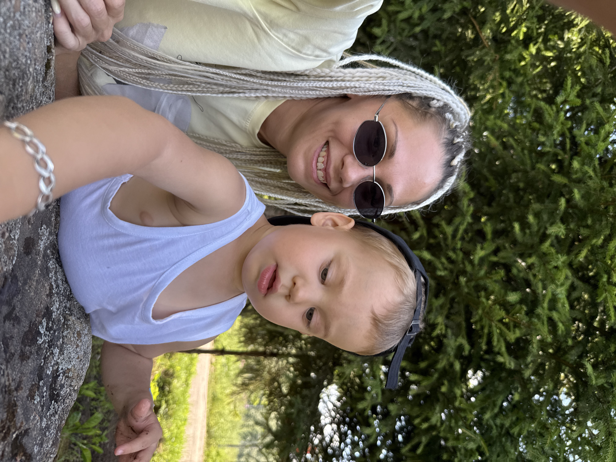 The media file shows a selfie of a woman and a young child outdoors. The woman, with blonde braided hair and sunglasses, is smiling at the camera. She is wearing a light yellow top. The child, in a white tank top and a black baseball cap, is looking towards the camera with parted lips. They appear to be close together, possibly leaning against a rock or tree. The background is filled with lush green pine trees, suggesting a natural, wooded setting. The lighting indicates it is daytime, likely sunny, with dappled sunlight filtering through the trees. The overall mood seems happy and relaxed, suggesting a casual outing. The location context is noted as Rusanovo, Russia.