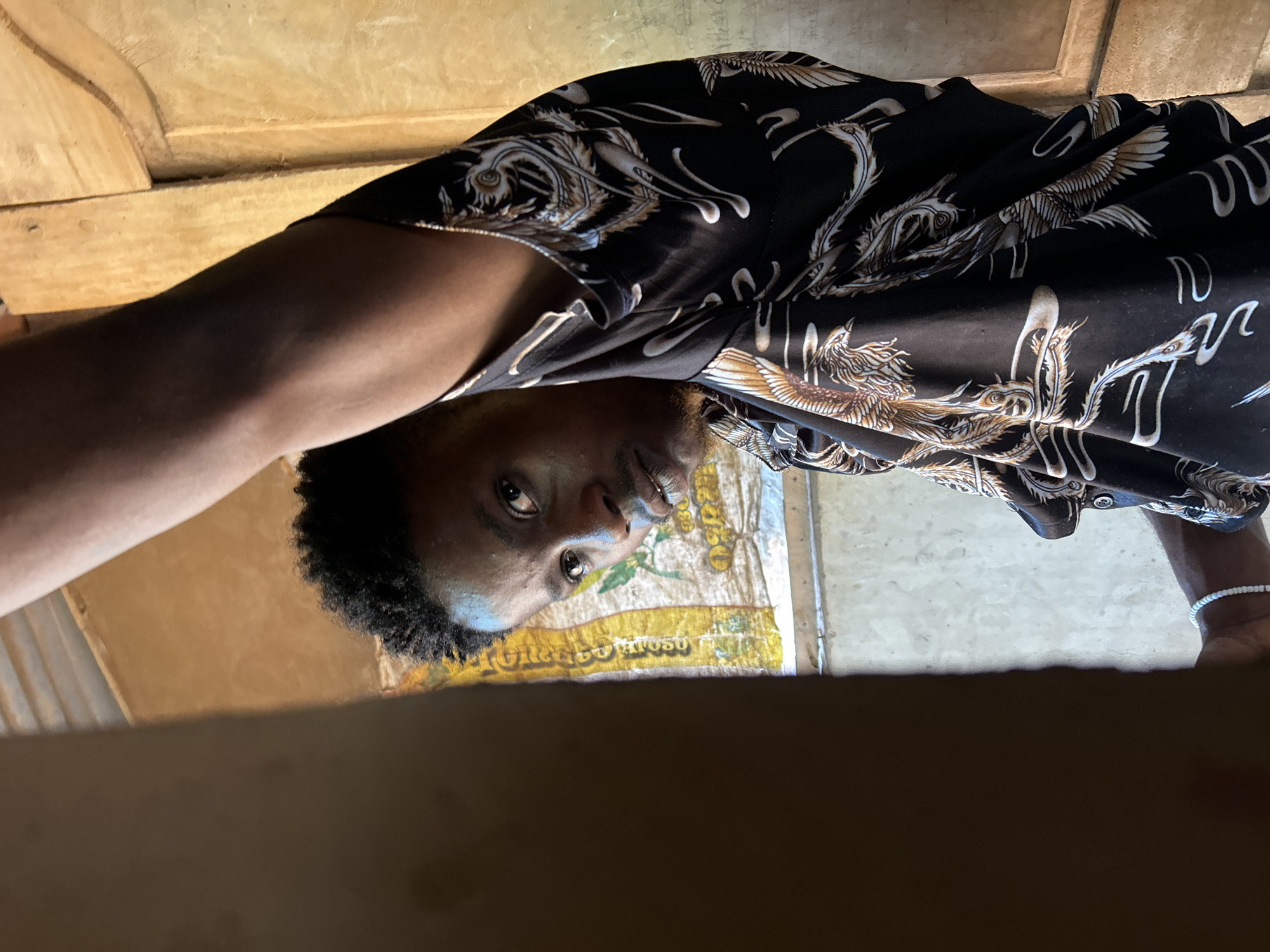 A low-angle selfie captures a person with dark skin, short curly hair, and stubble. They are wearing a black shirt patterned with ornate gold and white designs. Part of their bare arm and wrist, adorned with a white beaded bracelet, are visible. The person appears to be looking down at the camera. Behind the person is a yellow sack printed with the words "TORREON ATOSSO." The background consists of wooden surfaces.