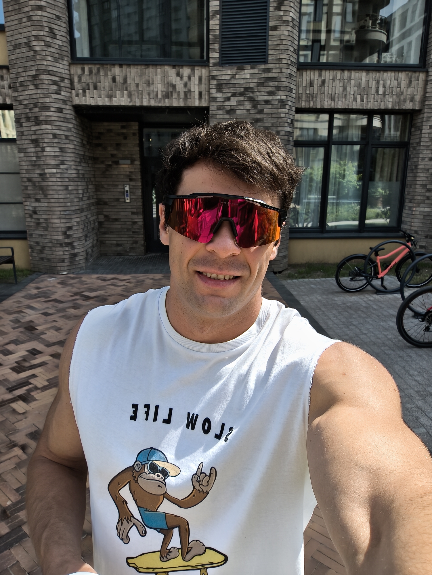 A man stands on a brick-paved area in front of a multi-story building with a modern brick facade and large windows, likely an apartment complex. He is wearing a white tank top with a graphic of a monkey on it, and reflective sunglasses. Two bicycles are parked to the right of the frame. The location is Moscow, Russia.