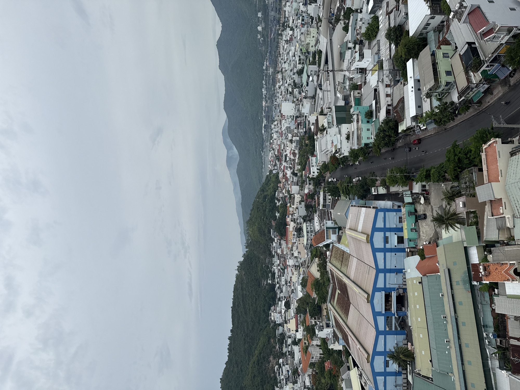 The image shows an aerial view of a coastal city, likely Nha Trang, Vietnam, with a dense urban landscape bordered by lush green hills and mountains in the background. The sky is overcast with a thick layer of grey clouds. In the foreground, a large, multi-sectioned warehouse-like building with a prominent blue and white facade dominates the left side of the frame. A road runs through the middle of the city, with several motorcycles and a few cars visible. The city itself is characterized by a mix of low-rise and mid-rise buildings, many with white or light-colored walls and various roof types, including terracotta tiles and corrugated metal. Vegetation is scattered throughout the urban area, with numerous trees and patches of green spaces. The background reveals a bay or body of water with a few boats, and further beyond, a range of mountains fading into the hazy distance. There is a subtle haze or light smoke rising from the mountain slopes in the background, suggesting potential agricultural burning or industrial activity. The scene depicts a typical day in a densely populated coastal city with surrounding natural topography.