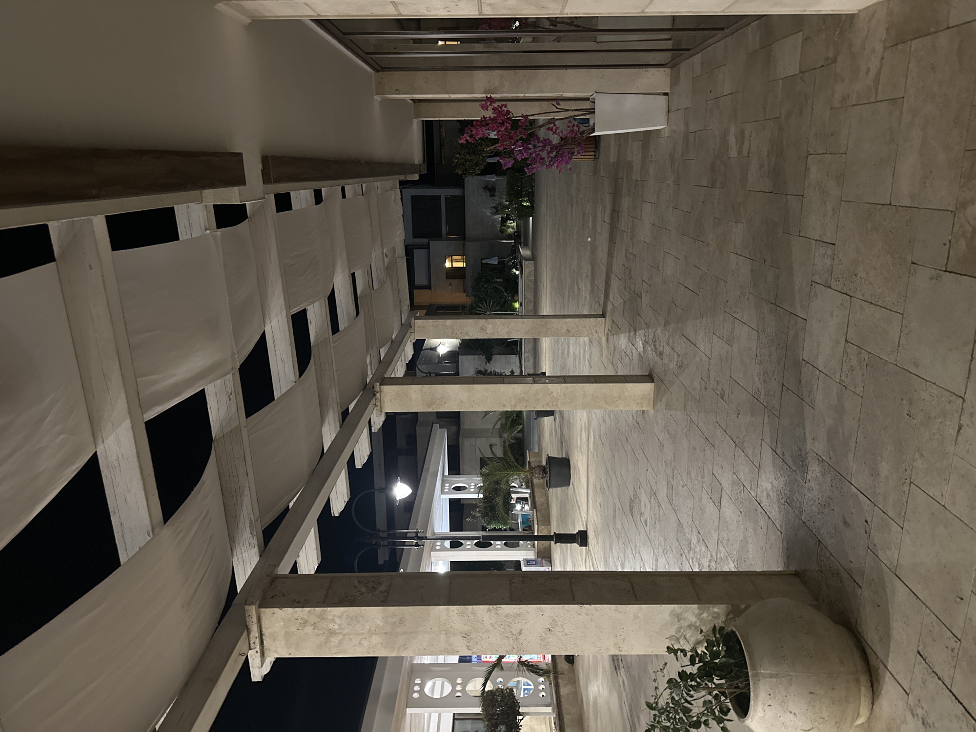 The image depicts an outdoor walkway at night, likely part of a hotel or resort complex in Sozopol, Bulgaria. The walkway is paved with light-colored rectangular tiles and is covered by a pergola-like structure with white fabric awnings stretched between wooden beams. Large, rough-textured stone columns support the structure, defining the pathway.
In the foreground to the left, a large, round, off-white ceramic pot contains a small green plant. Further down the walkway, a tall black lamppost with a white globe light illuminates the area. To the right, a white rectangular planter holds a vibrant pink flowering plant. The background reveals a building with dark windows and a soft yellow light emanating from one of them, suggesting an interior space. Landscaped gardens with palm trees and other greenery are visible in the middle ground. The overall atmosphere is calm and serene, typical of an evening setting in a tourist destination. There are no people or animals visible in the scene.