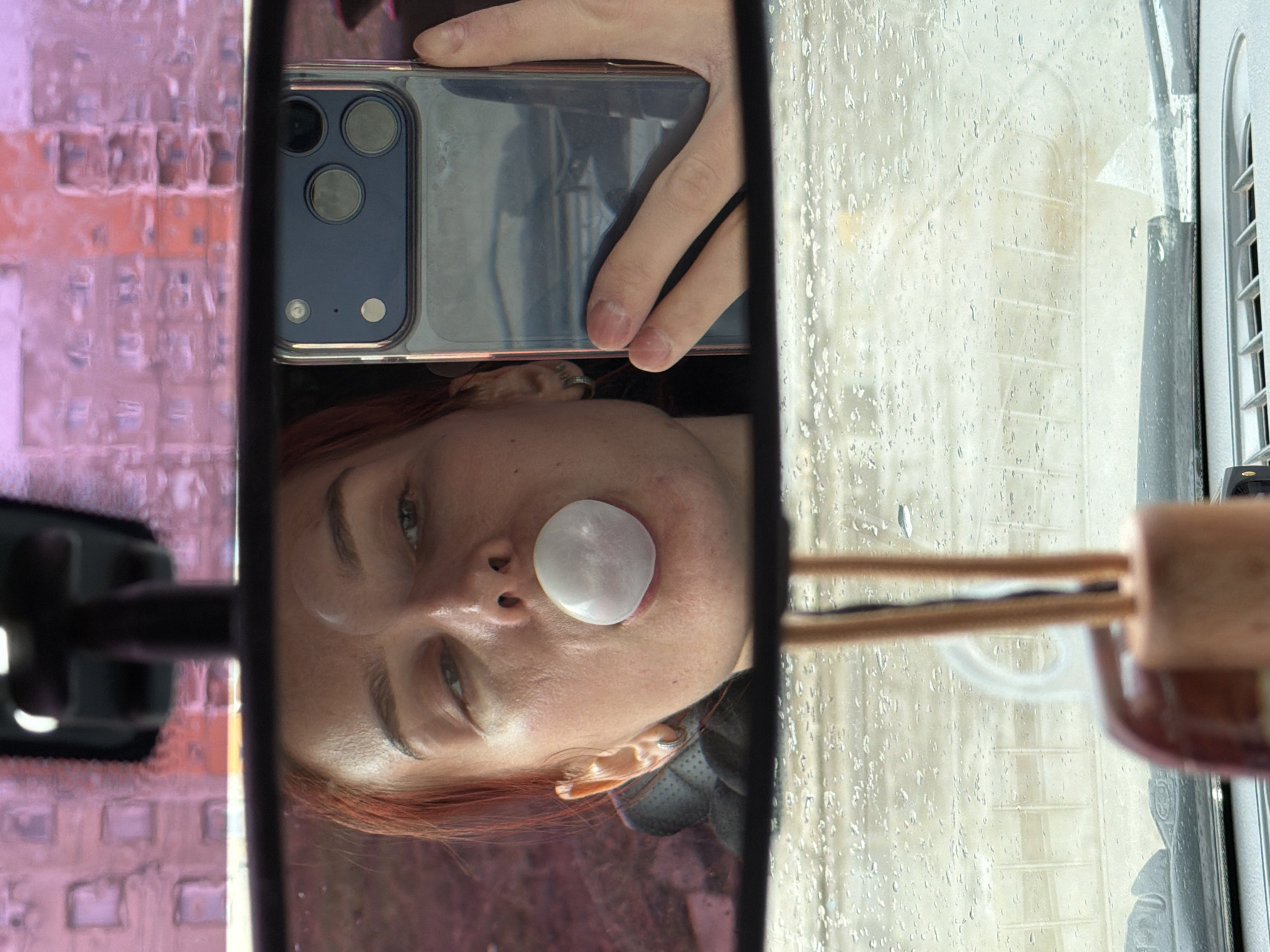 A selfie of a Caucasian woman with red hair is reflected in a car's rearview mirror. The woman has a large, white bubblegum bubble covering her mouth and nose. She is holding an iPhone in her left hand, the phone's camera lens facing the mirror. The background seen through the windshield is blurred due to raindrops. On the left side of the mirror's reflection is an out-of-focus structure, possibly a building, with a pinkish hue. On the right side, the car's window and frame are visible. An air freshener is attached to the mirror on the right side. The scene suggests the selfie was taken inside a car on a rainy day. The location in the city of Chaikovsky, Russia, cannot be determined from this image alone.
