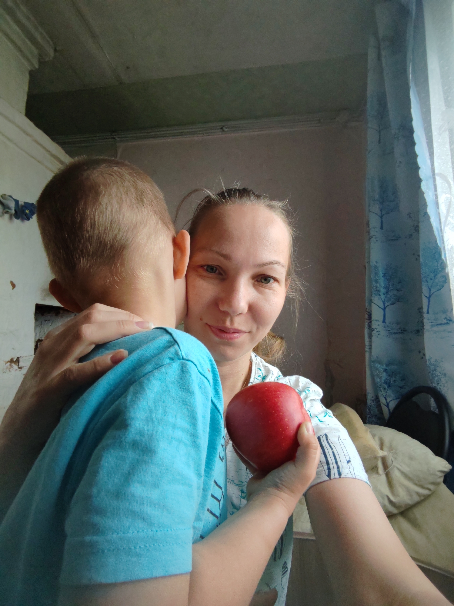 A woman in Чухлэм, Russia, holds a red apple in her right hand. Her left arm is around a child with short blonde hair. The child is wearing a blue t-shirt. Behind the woman are a window with patterned curtains, a pillow, and a chair. In the background on the left is a white wall.