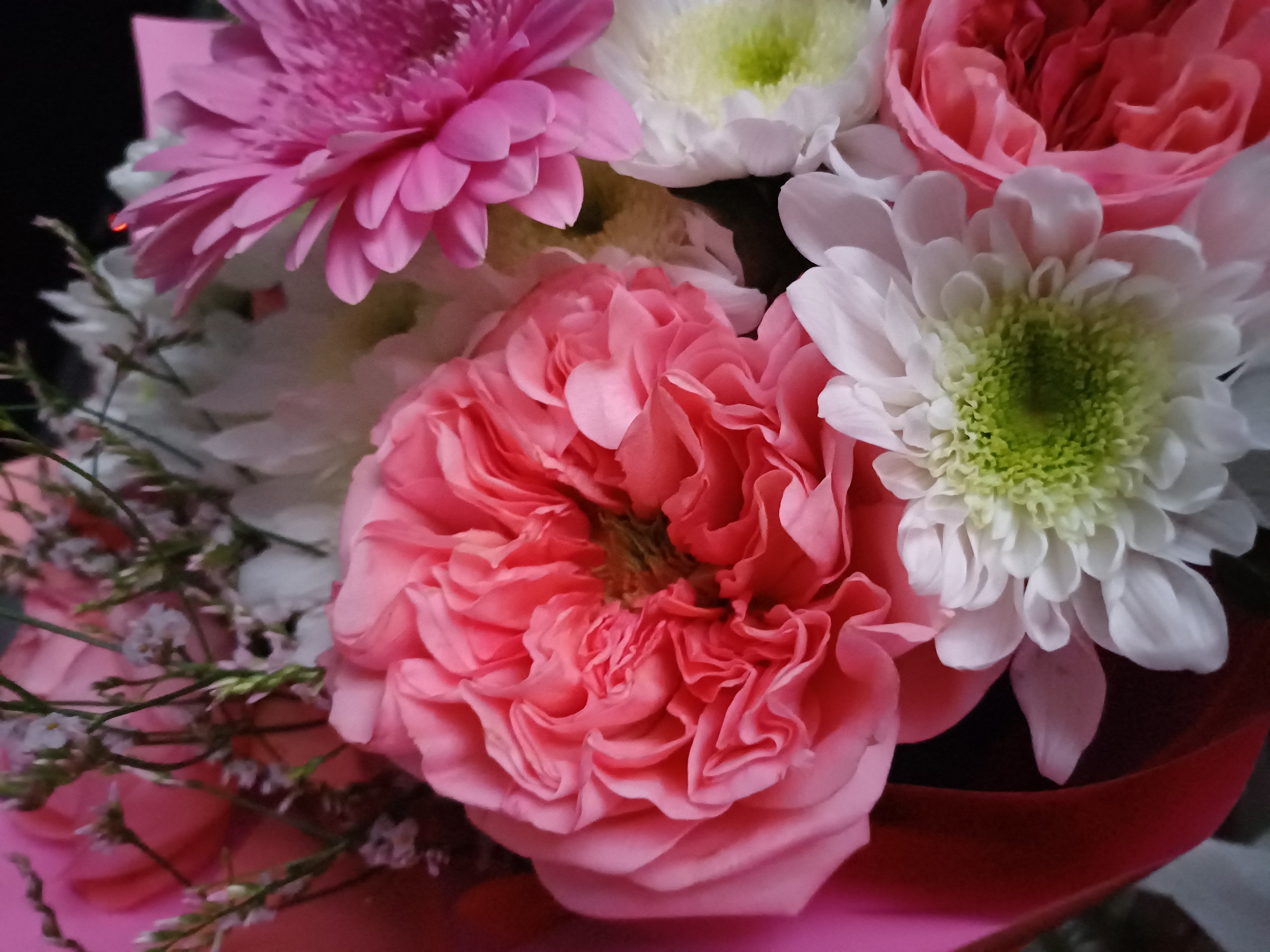 The image is a close-up shot of a bouquet of flowers. The bouquet features several different types of flowers in shades of pink and white, with some delicate greenery and smaller, pale purple flowers interspersed. Dominating the center is a large, intricately petaled pink rose. To its right, a bright pink daisy-like flower is visible, and below it are white chrysanthemum-like flowers with light green centers. A bright pink wrapping paper peeks out from the left side of the frame, hinting at the bouquet's presentation. The lighting is soft, highlighting the textures and colors of the flowers. There are no people or environmental details of a city visible.