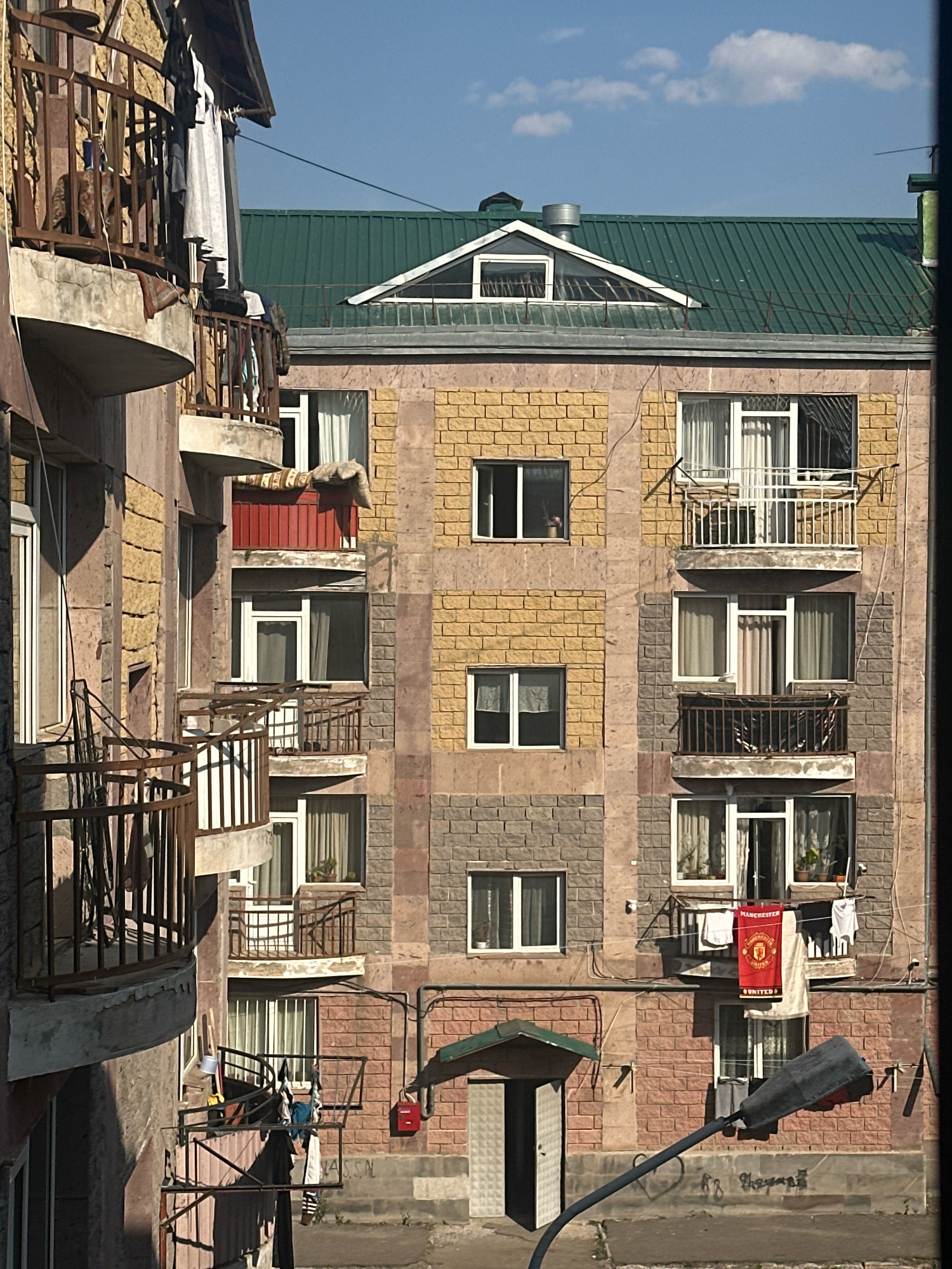 This is a medium-shot, eye-level photograph taken in Gyumri, Armenia. The scene depicts the exterior of an apartment building with balconies, windows, and a green metal roof. The building's facade is constructed with a combination of light yellow and gray brickwork. Several balconies are visible, adorned with railings and some with laundry hanging to dry. One balcony on the right side of the building displays a red banner with a crest and the words "MANCHESTER UNITED". Multiple windows are open or closed, with curtains visible from some. A street lamp with a curved arm is positioned in the foreground, extending into the lower right of the frame. The sky is blue with scattered clouds. The overall impression is of a residential urban environment.