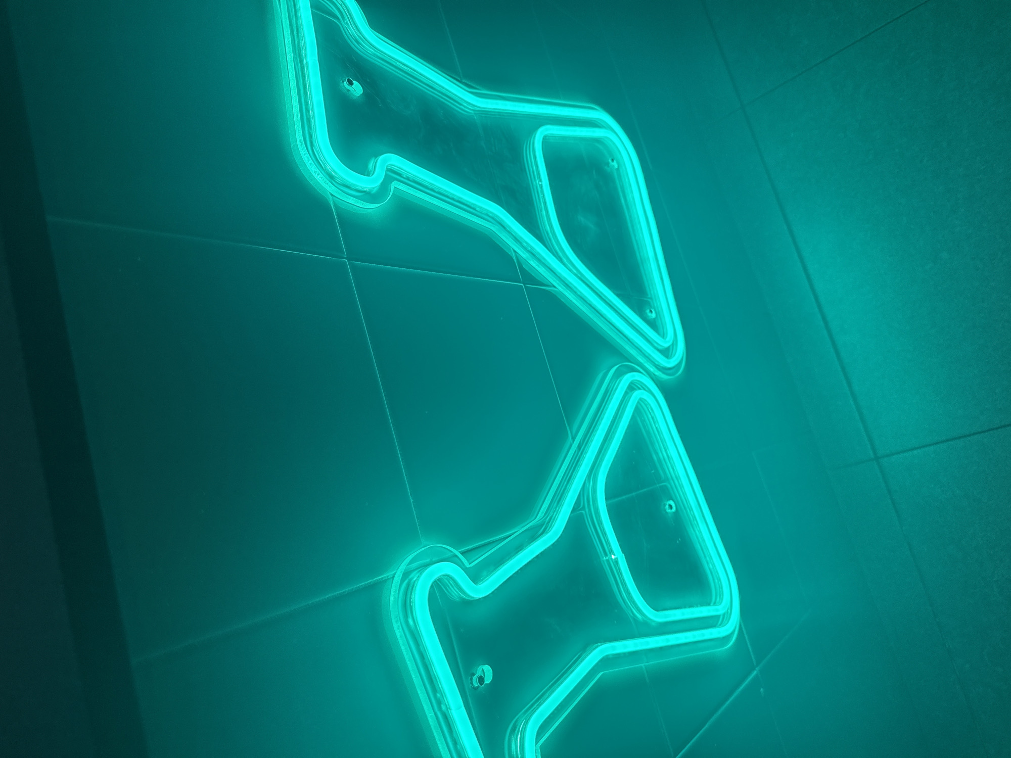 Two bright teal neon shapes, resembling stylized megaphones or racetrack outlines, are mounted on a tile wall. The tiles are a matching teal color with grout lines visible. The image is taken at a low angle, enhancing the three-dimensional aspect of the neon signs. There is no other contextual information that indicates this is in San Bernardino Tlaxcalancingo, Mexico.