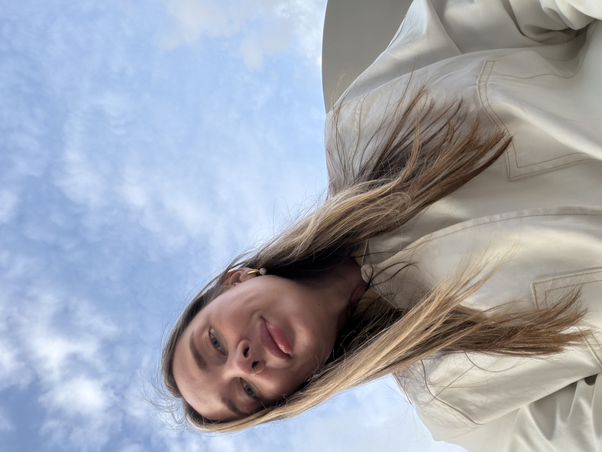 A woman is photographed from a low angle against a backdrop of a partly cloudy blue sky. Her face is partially visible, displaying blue eyes, a nose piercing, and light-colored hair. She wears a white garment. The location cannot be determined from the provided image, so the reference to Moscow, Russia, cannot be confirmed.