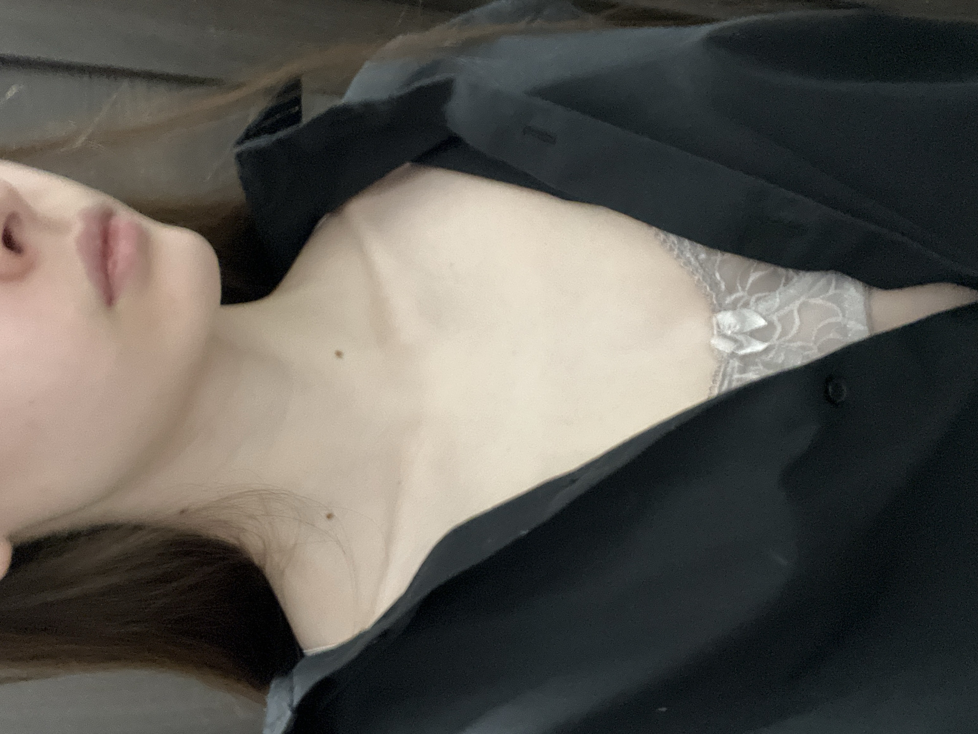The image shows a person lying down, wearing a black shirt that is partially unbuttoned, revealing a white lace garment underneath. The person's face is partially visible, showing the nose and mouth. The location is not discernible from the image.