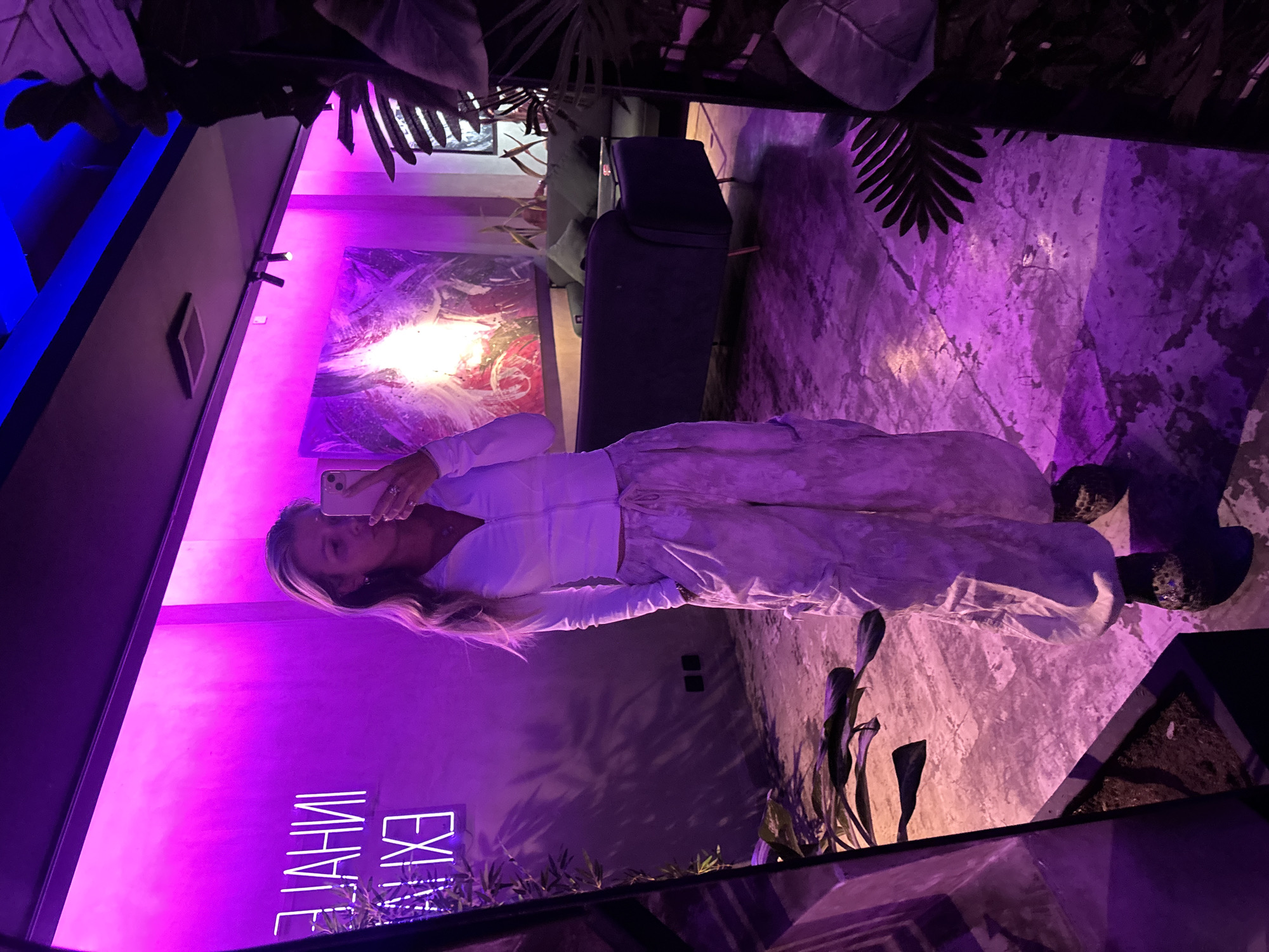 A young woman stands in front of a large mirror, taking a selfie. She is wearing a white long-sleeved cropped zip-up shirt and baggy light-colored cargo pants. Her hair is long and blonde, styled in loose waves. In the background, reflected in the mirror, is a wall with neon signs that read "INHALE" and "EXHALE" in white lettering. There is also a large abstract painting with vibrant colors, a dark sofa, and several potted plants. The room is illuminated by purple and blue lights, creating a moody ambiance. The floor appears to be concrete. The overall scene suggests an indoor space, possibly a lounge or a studio.