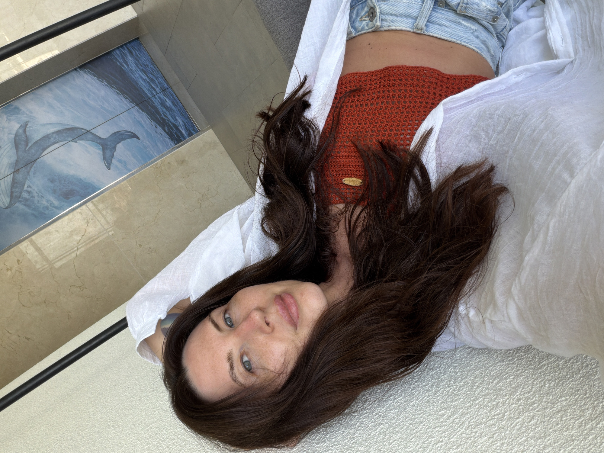 A woman with long, dark brown hair and blue eyes is lying down, looking at the camera. She is wearing a white unbuttoned shirt, an orange crocheted top, and light-wash denim shorts. Her left arm is resting behind her head. The background features a textured wall on the left and a large framed artwork of a whale in the ocean on the right. The overall environment appears to be indoors, possibly a hotel room or a modern living space, given the artwork and marble-like wall finish. The image does not contain any specific visual cues that would indicate the location as Dong Ninh Hoa Ward, Vietnam.